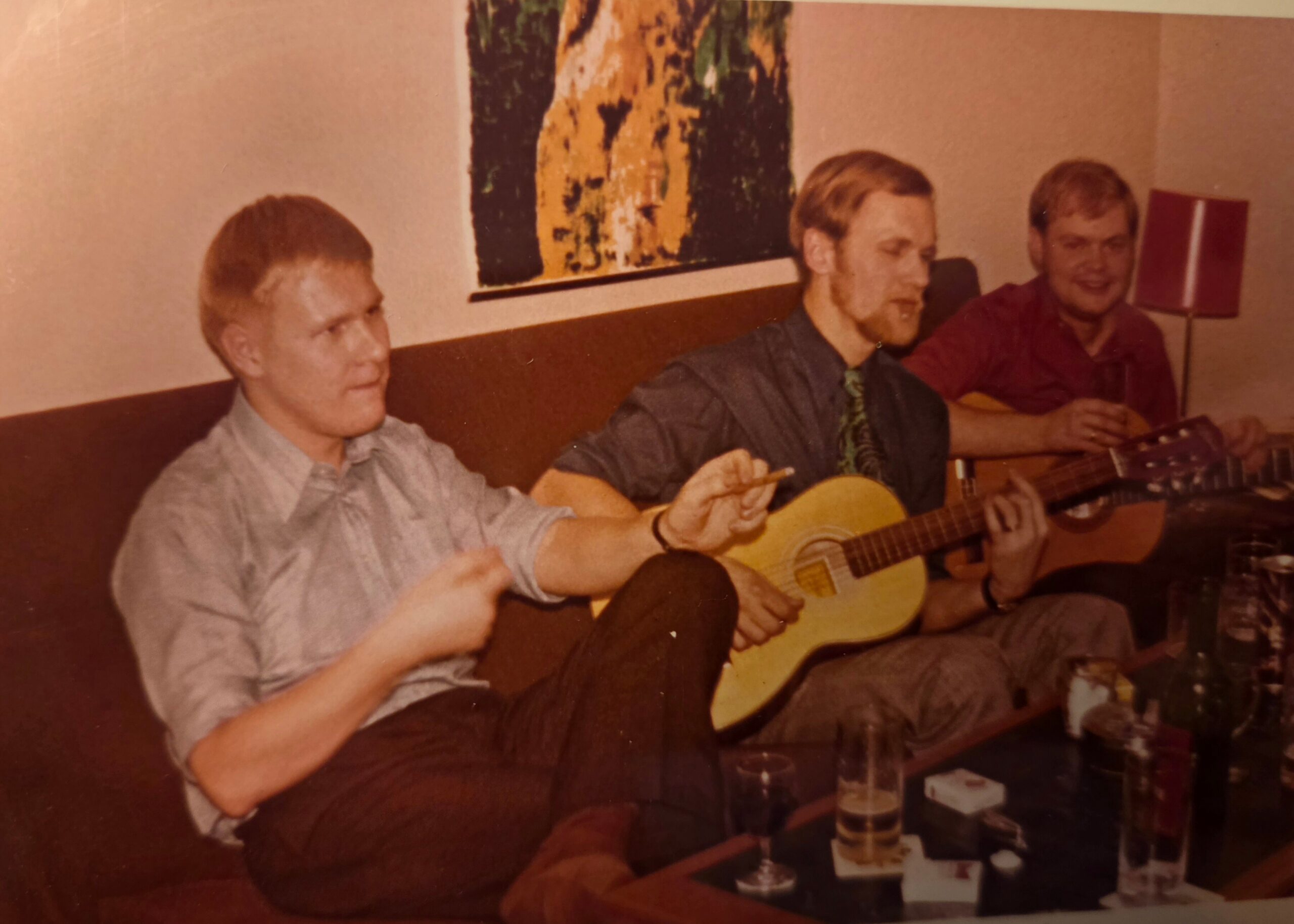 Three men sit on a couch playing acoustic guitars together in a cozy living room, with drinks on the table nearby (vintage photo).