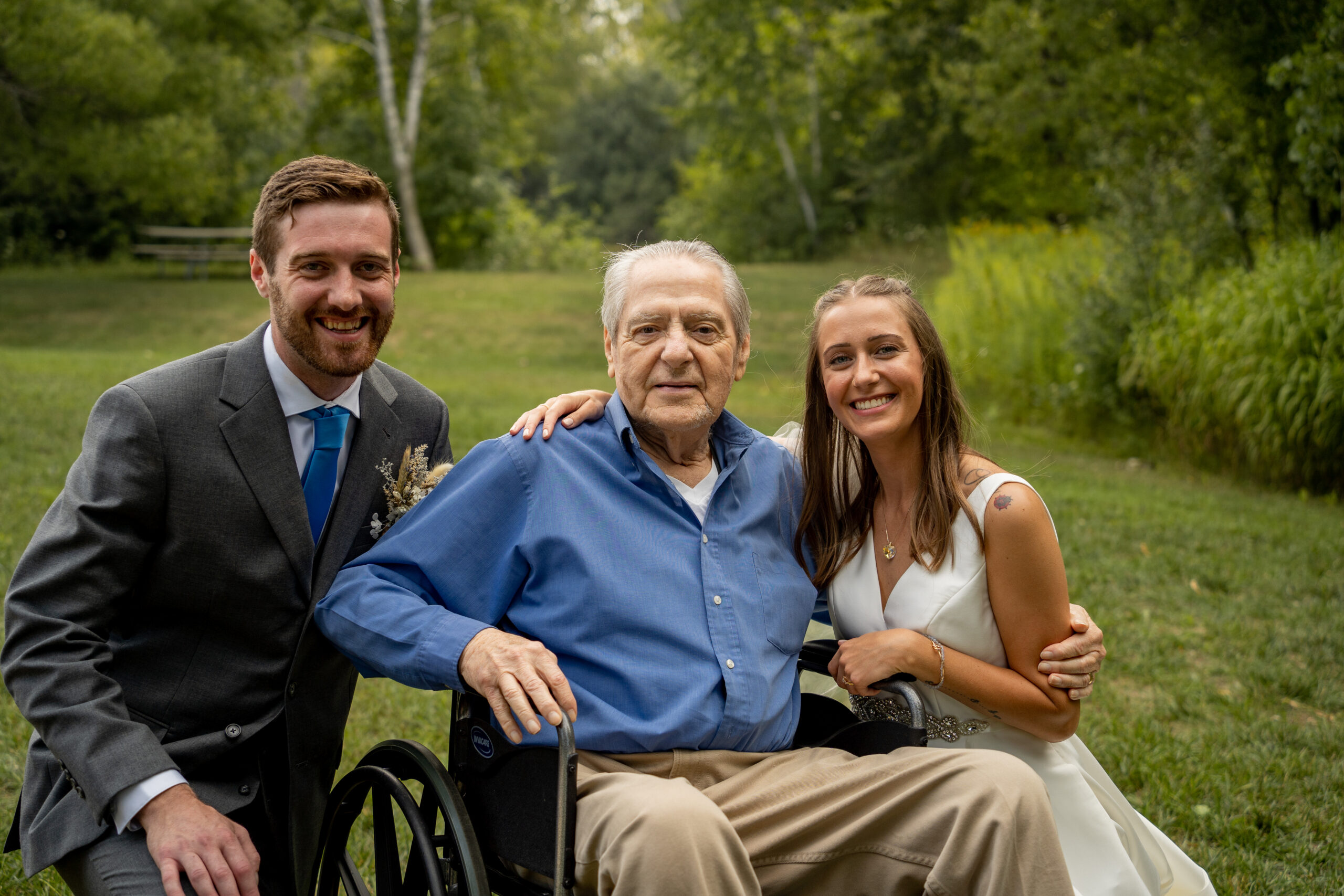 Three people posing outdoors in a park: a man in a gray suit, an older man in a wheelchair, and a smiling woman in a white dress.