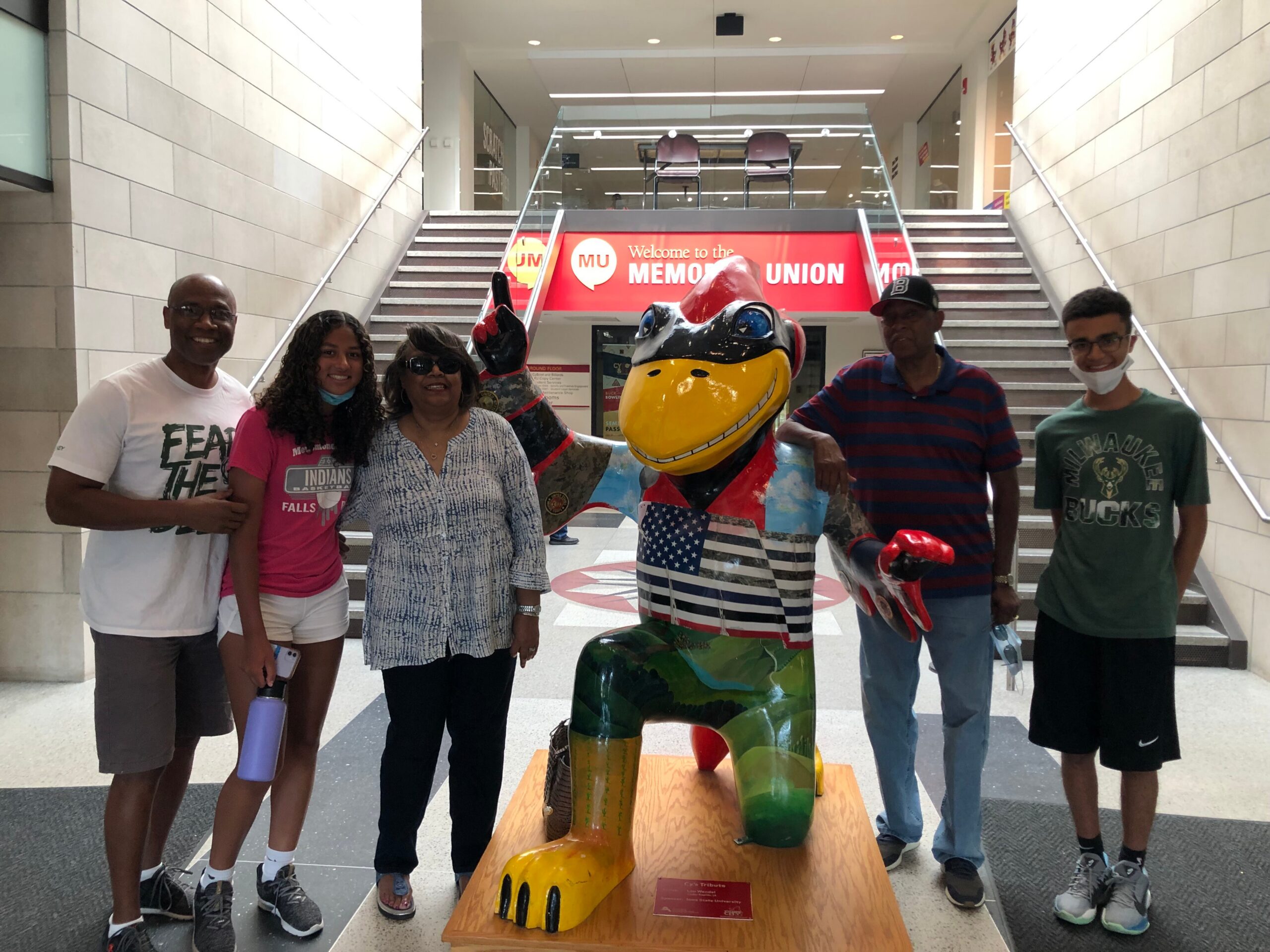 Six people pose with a large colorful sculptural figure in a bright, modern indoor space; a red banner reads 'Welcome to the Memorial Union' in the background.