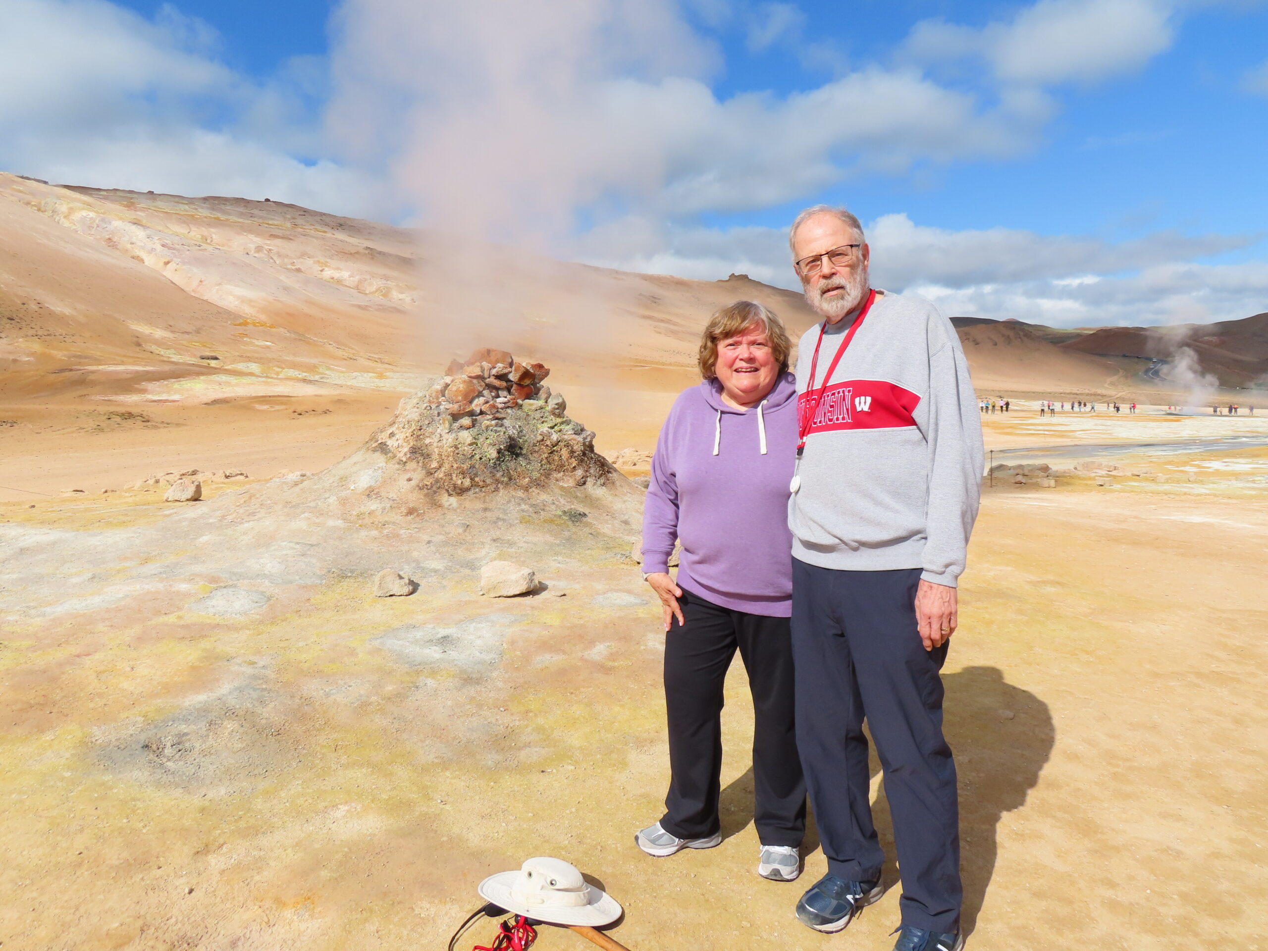 Two seniors stand together in a geothermal landscape with steam vents and a blue sky behind them.