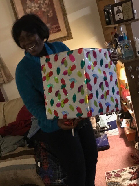 Smiling woman in a teal sweater holds a large wrapped present with a multicolored leaf pattern in a cozy living room.