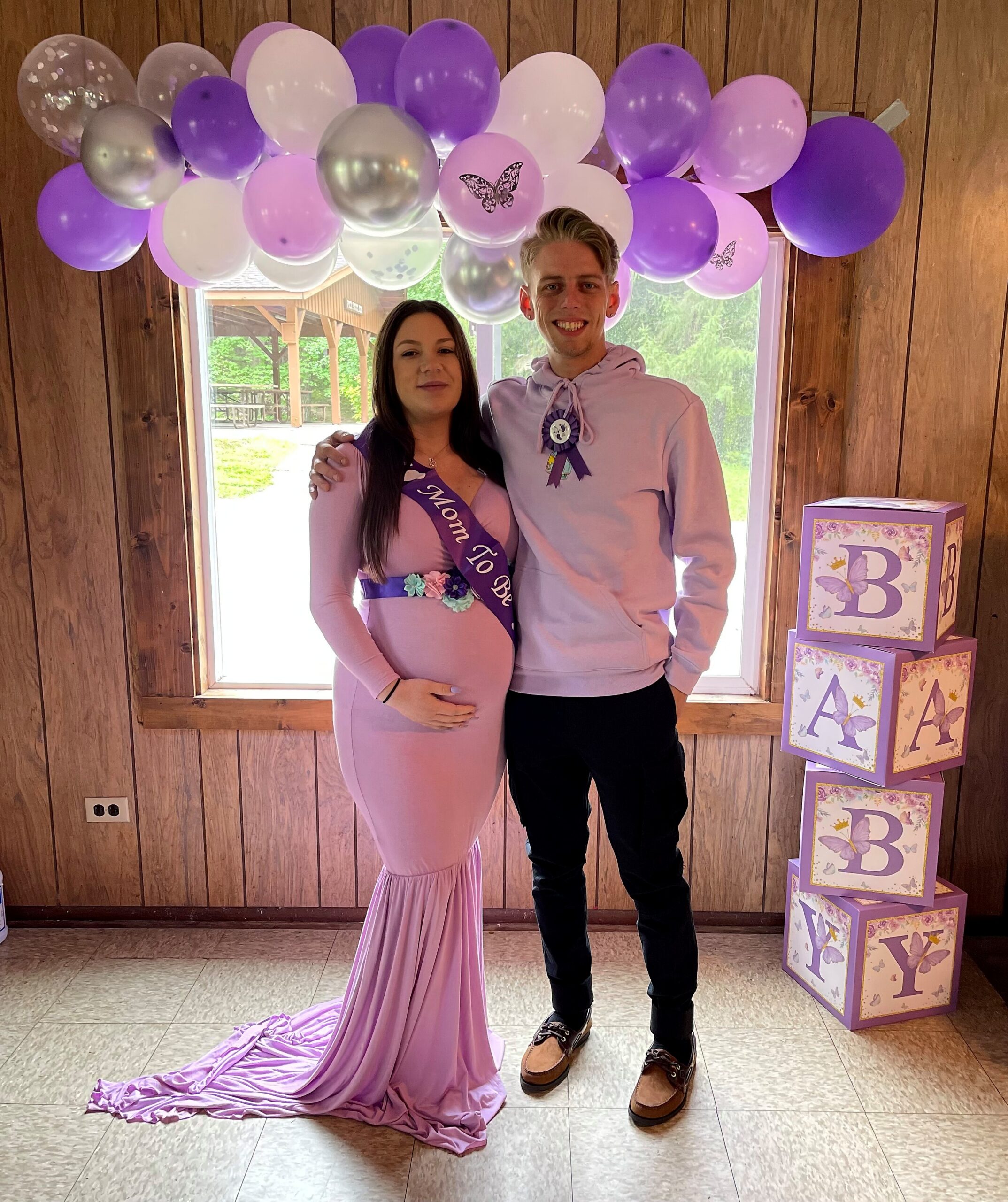 Pregnant woman in a pink gown with a 'Mom To Be' sash and a man in a lavender hoodie, posing under pastel balloon arch in a wood-paneled room.