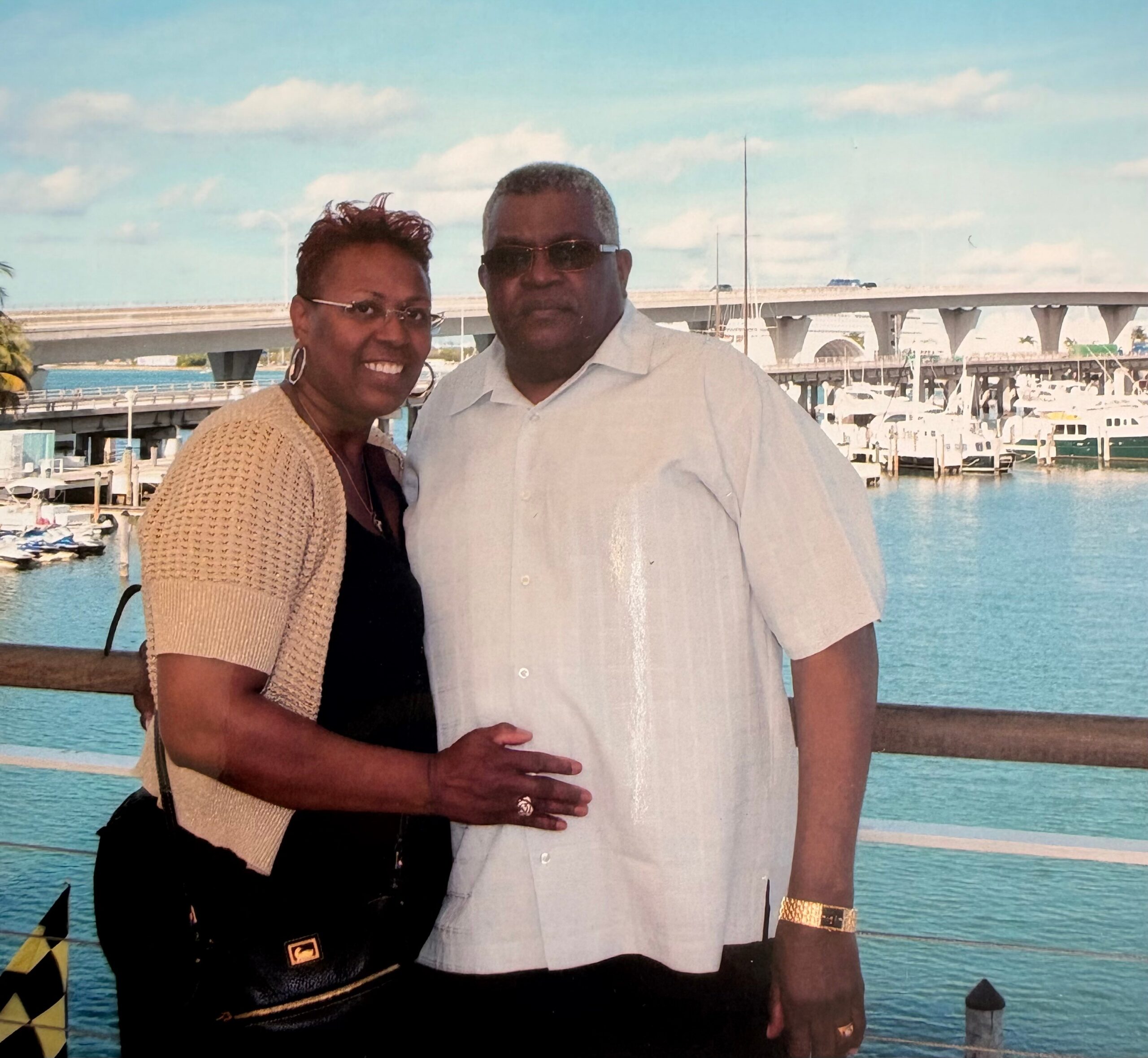Smiling couple standing close together at a marina, with boats and a clear blue sky behind them.