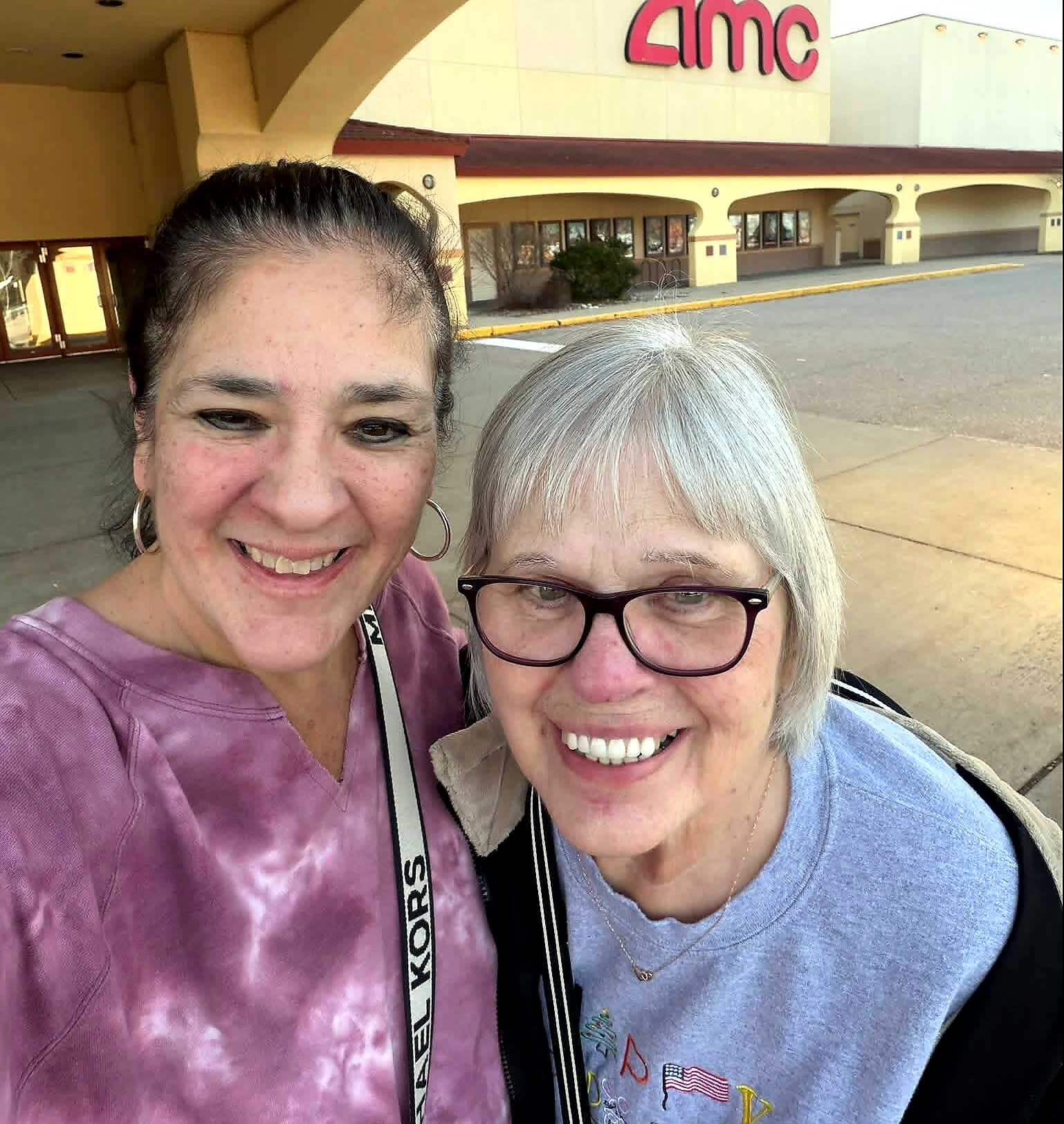 Two smiling women pose for a selfie outside an AMC theater storefront.