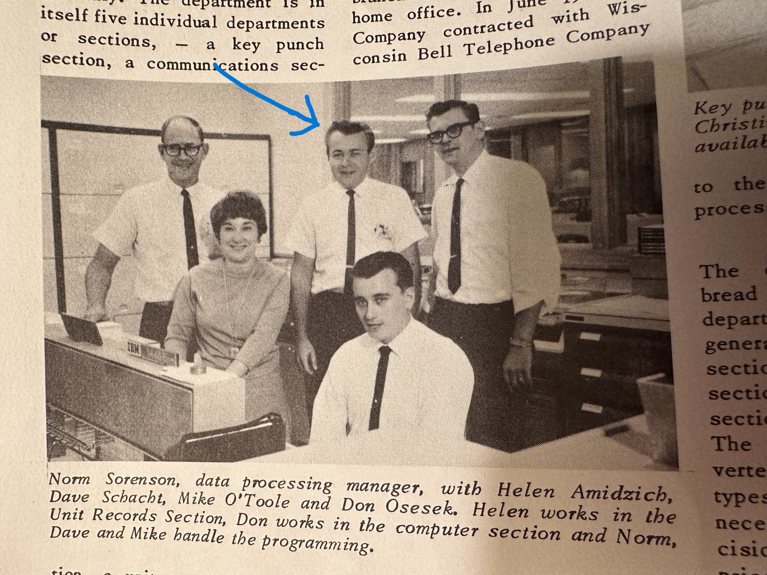 Group of six office workers in 1960s attire posing around early computer workstations in a lab.
