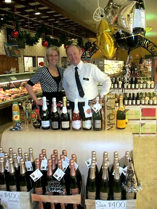 Two store staff pose behind a table of champagne bottles, with balloons overhead in a shop.