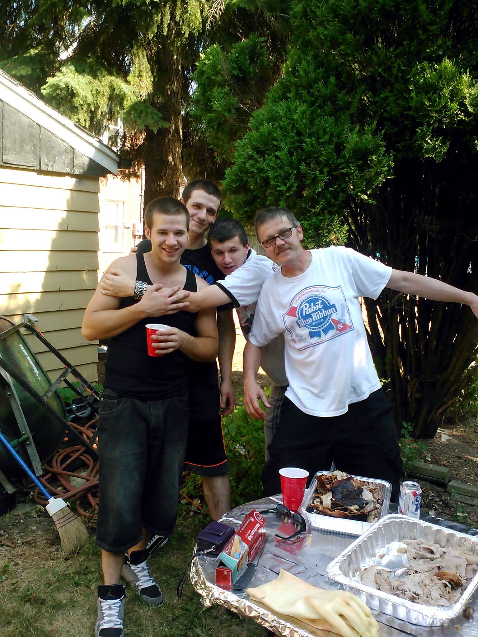 Four men posing together in a sunny backyard barbecue, with foil trays and red cups of food nearby.