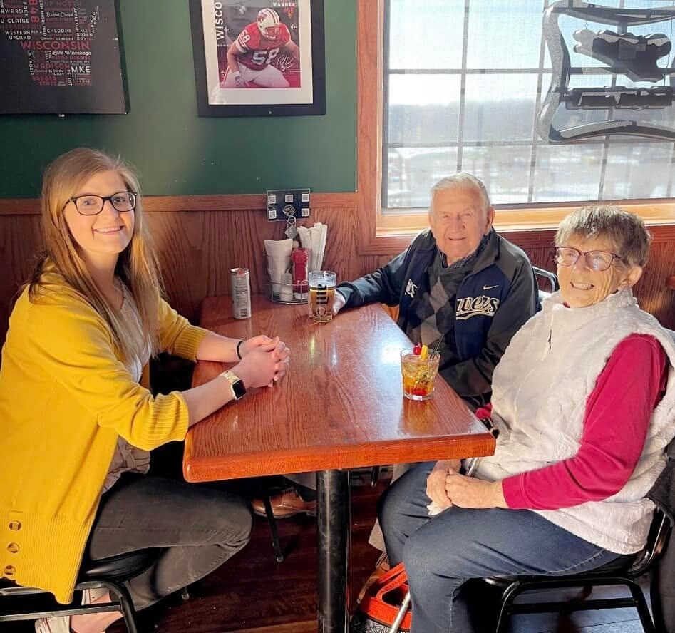 Three people sit at a wooden table in a café: a smiling young woman in a yellow cardigan on the left and an older couple on the right, with drinks on the table and wall art behind them.
