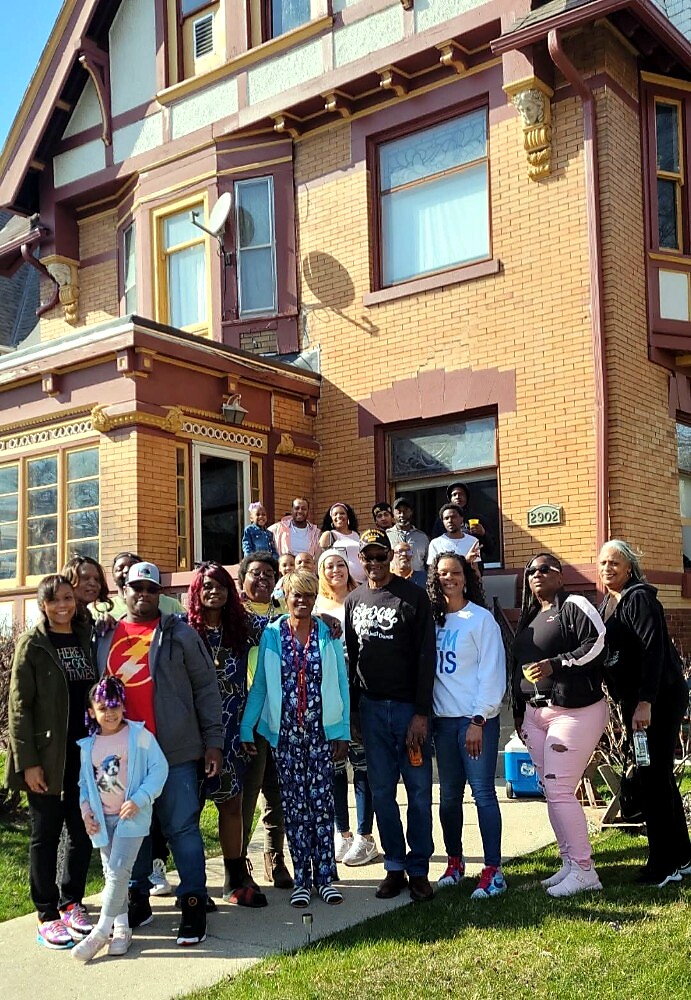 Diverse group of people posing for a photo on a sidewalk in front of a yellow-brick house with decorative trim.