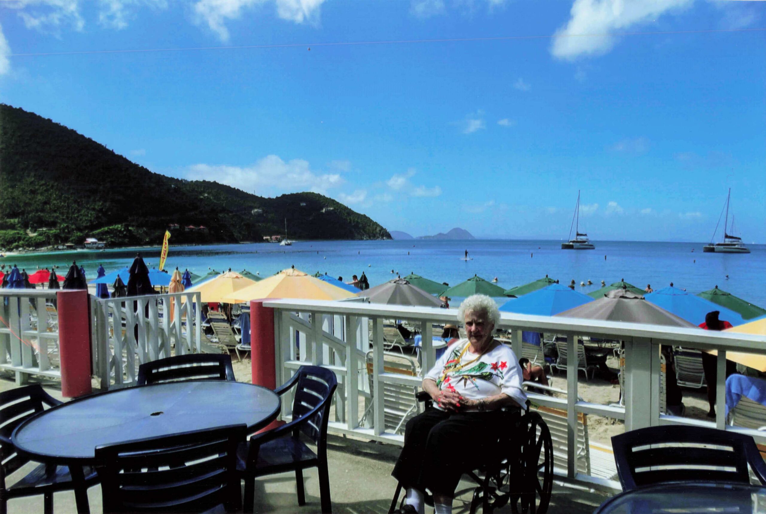 Beachside cafe area with colorful umbrellas, metal tables and chairs, and a man in a wheelchair seated at the railing in the foreground, ocean and hills in the background