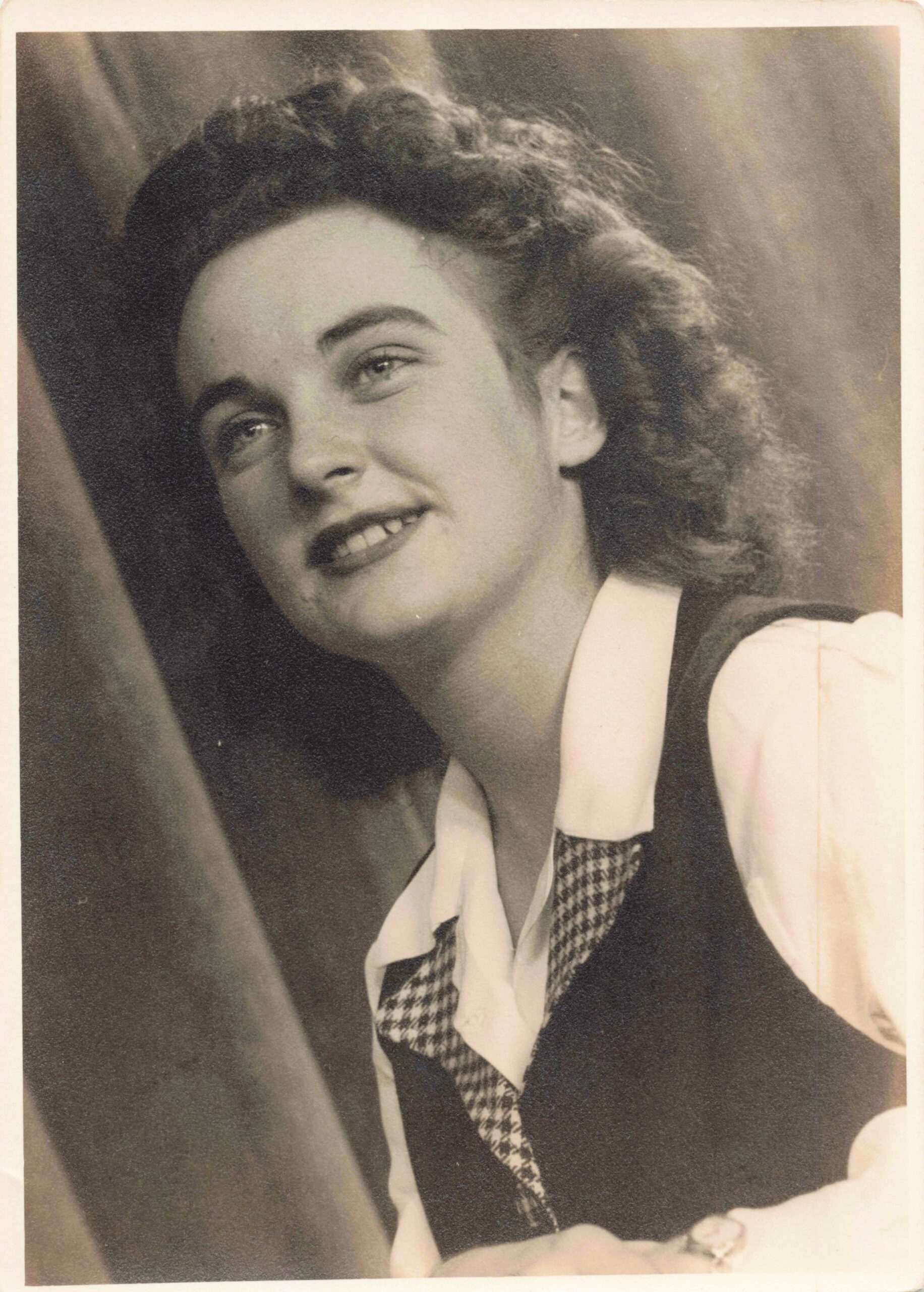 Young woman with wavy hair smiling, wearing a white shirt and houndstooth vest, in a vintage photo.