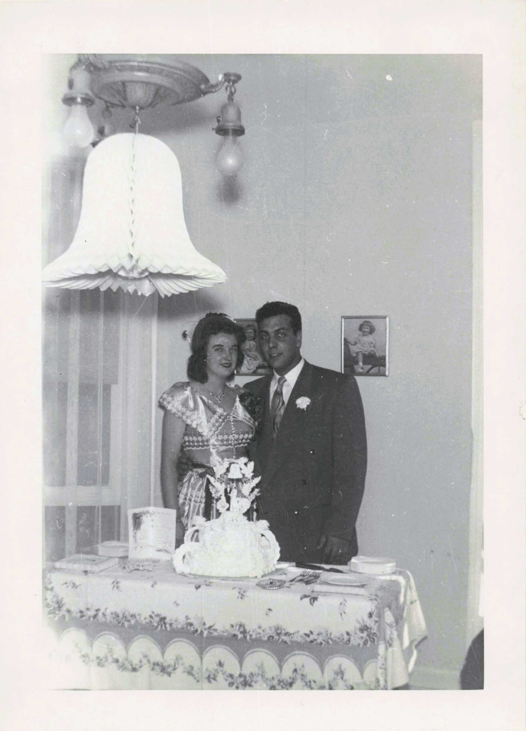 Couple in formal attire posing beside a decorated wedding cake on a table, with a hanging chandelier above.