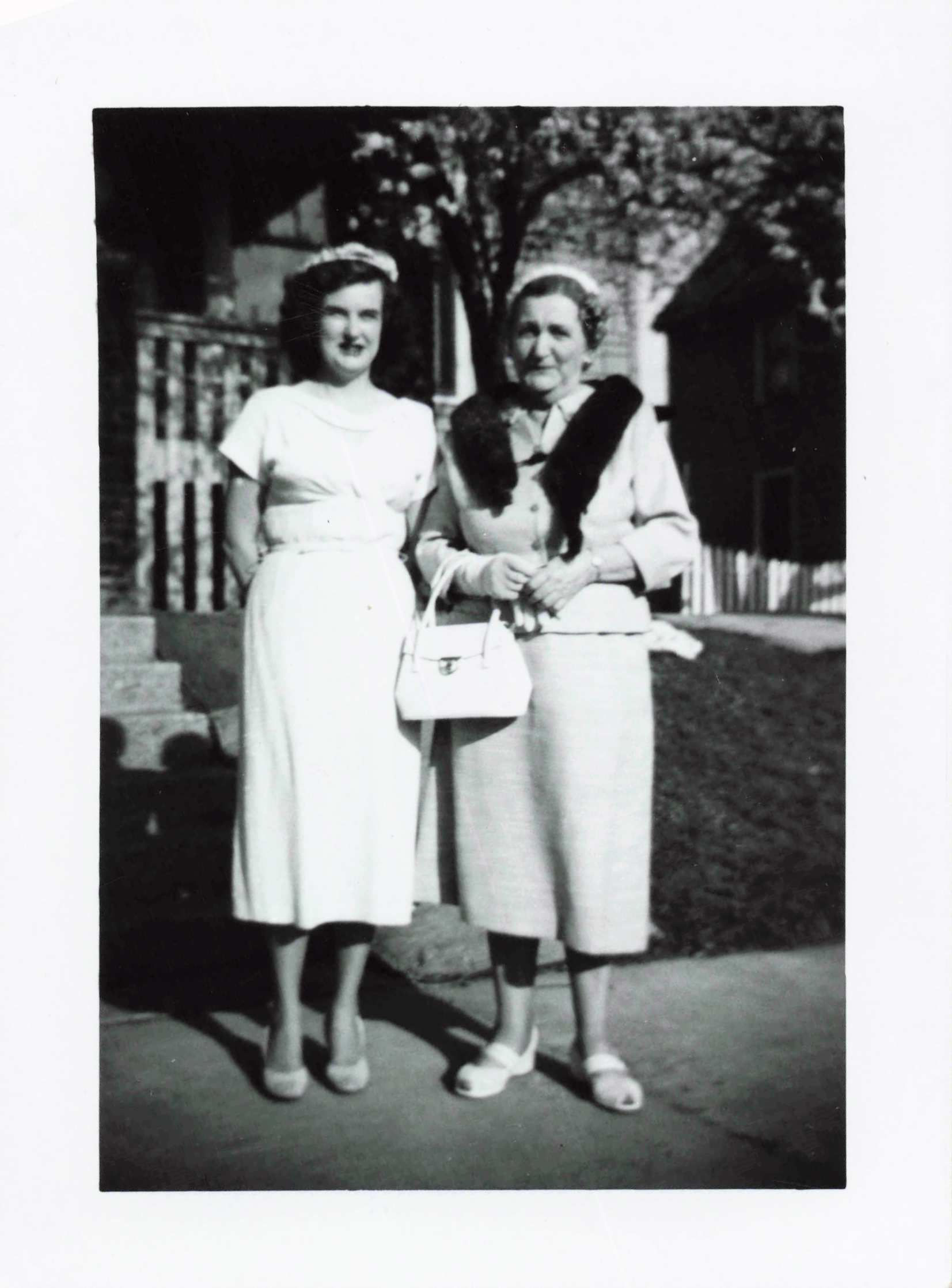 Two women stand side by side outdoors in vintage dresses; the younger left wears a white dress and headband, holding a small handbag, while the older right wears a light coat with a fur stole and headband.