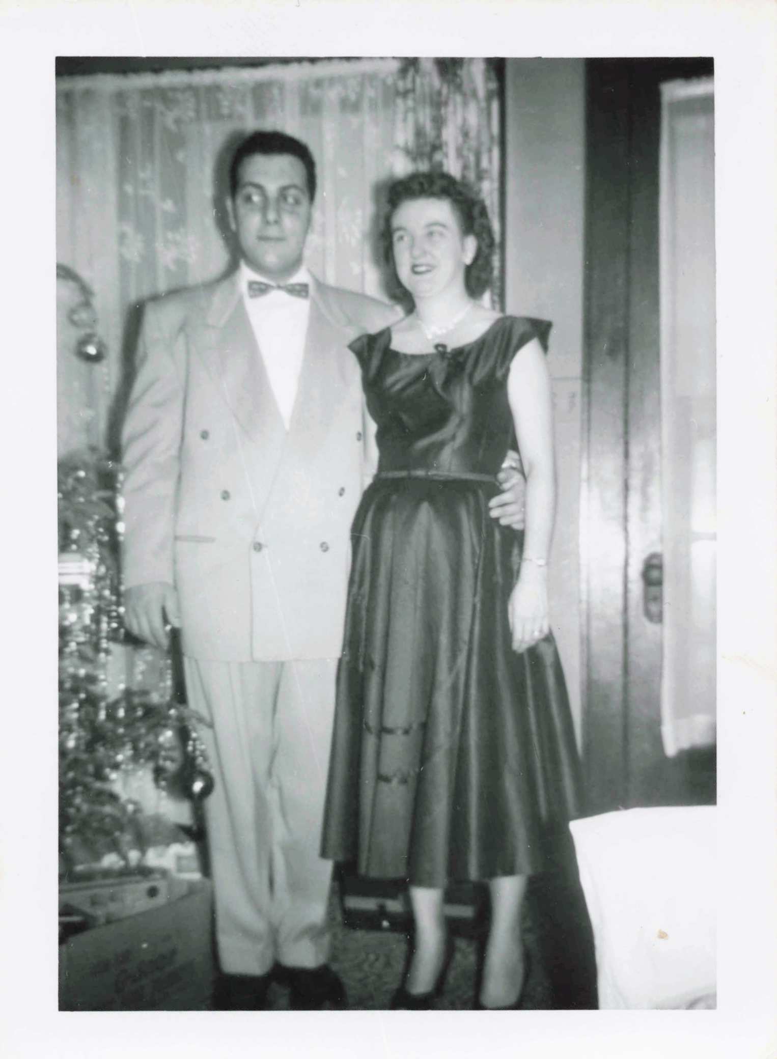 Man in a light-colored suit with a bow tie standing beside a woman in a dark satin evening gown; they smile together indoors.