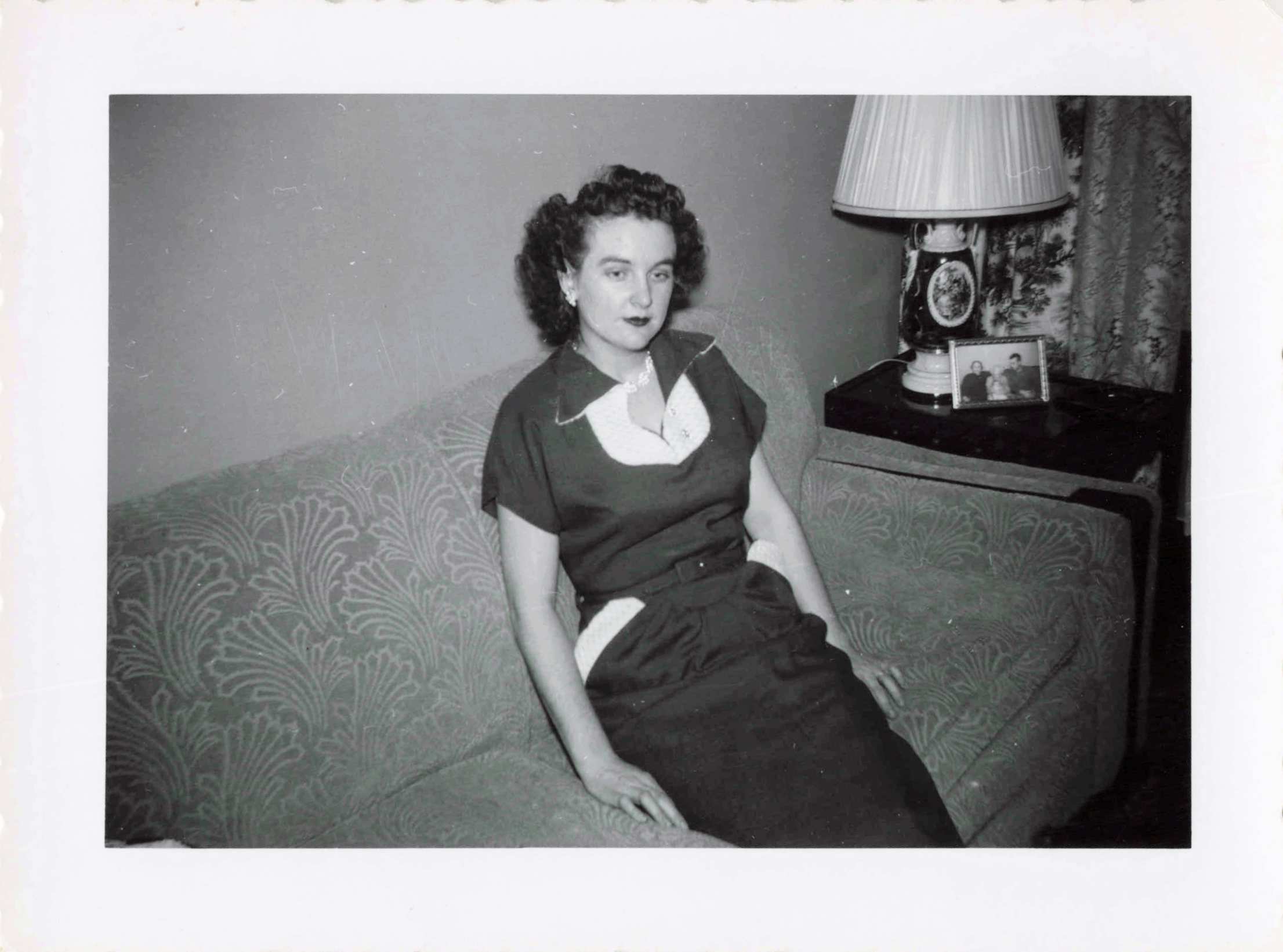 Black-and-white photo of a woman in a vintage dress sitting on a patterned sofa, with a ornate lamp and framed photograph on a side table beside her.