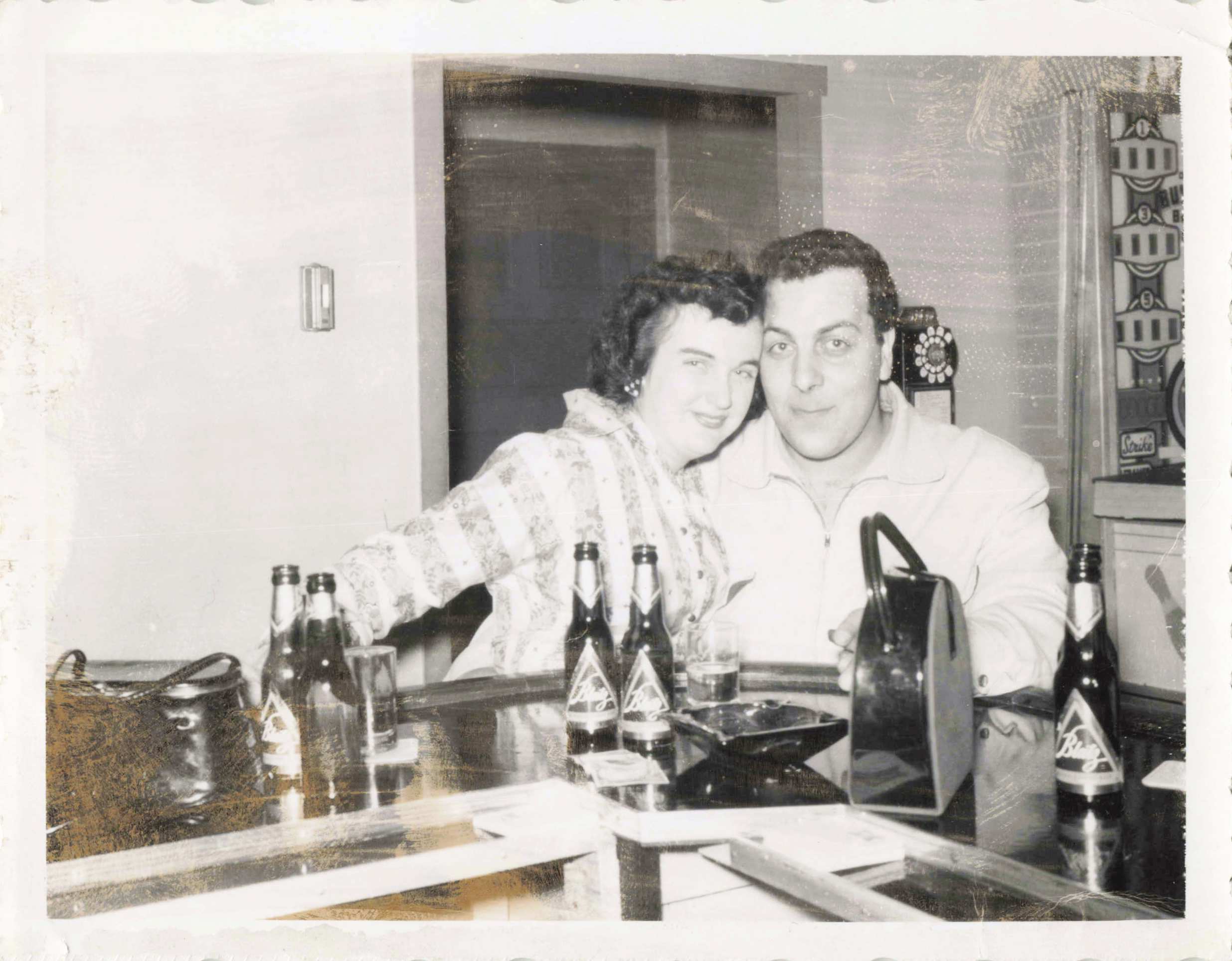 Smiling couple leaning close together at a table with soda bottles in front, posing for a casual snapshot in a home setting