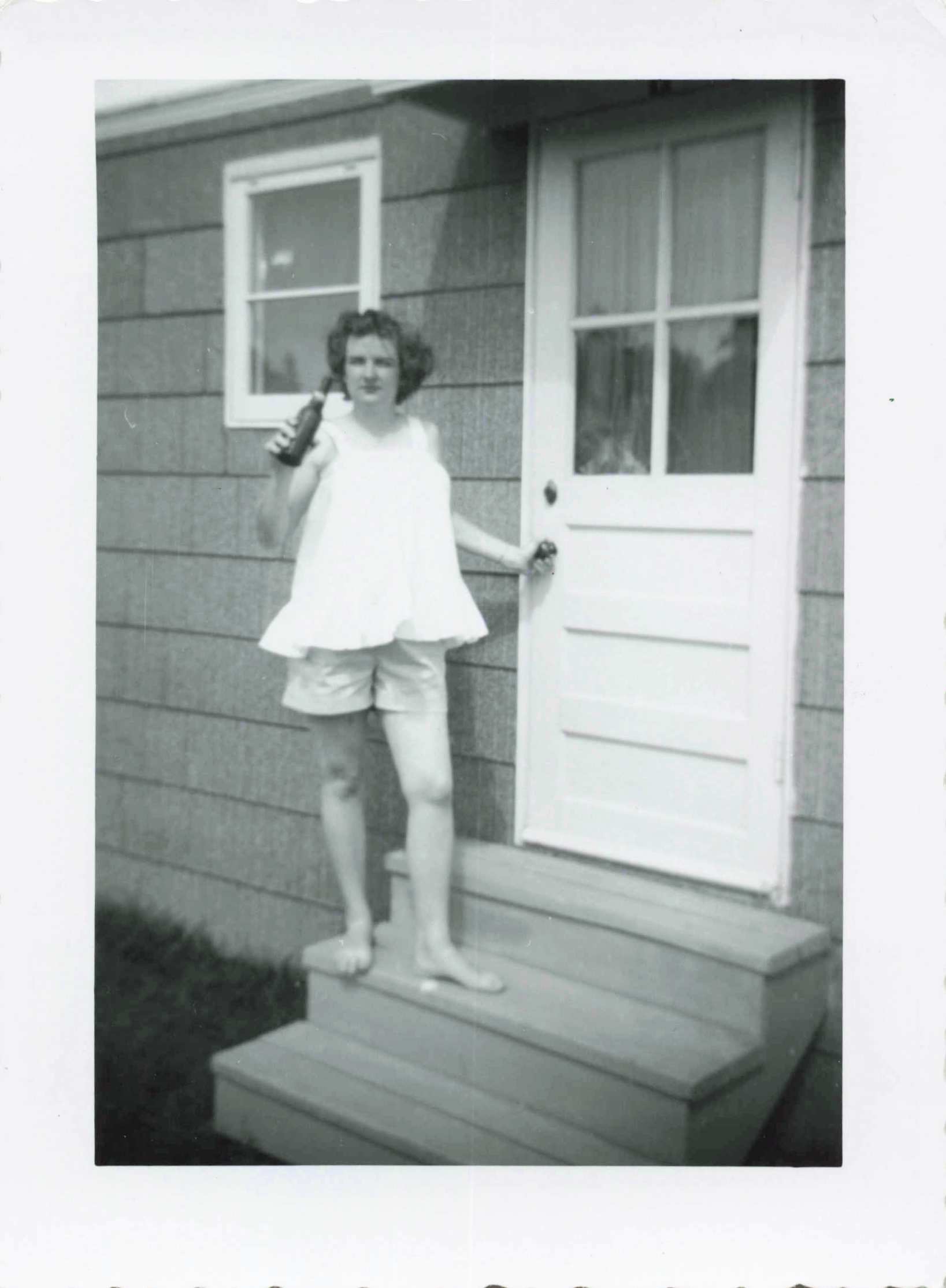 Young woman stands on wooden front steps outside a house, holding a bottle in her right hand and touching the door with her left hand, wearing a light sleeveless top and shorts (black-and-white photo).