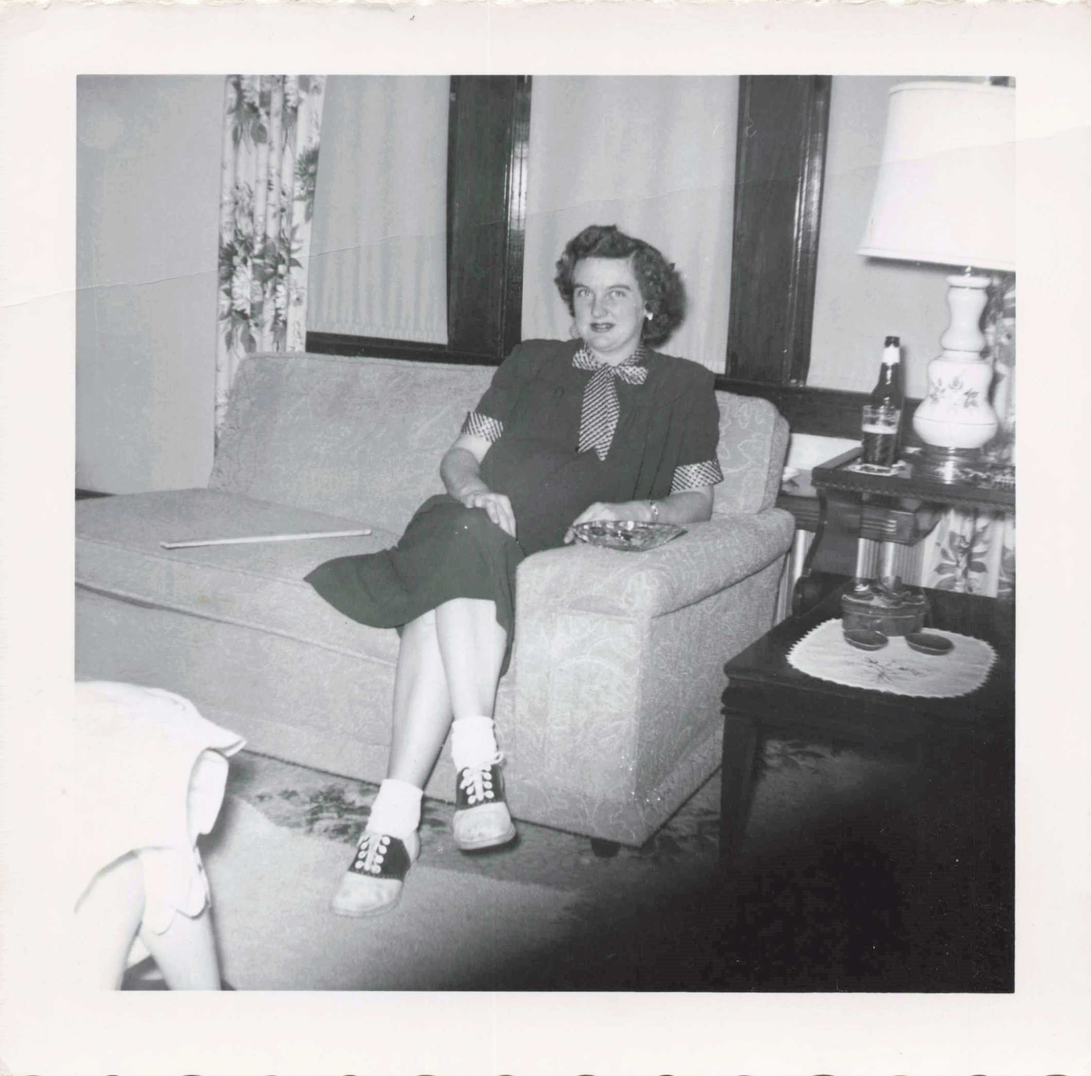 Black-and-white photo of a woman sitting on a sofa in a living room, wearing a dark dress with a checkered scarf, hands folded in her lap, with a lamp on a side table nearby.