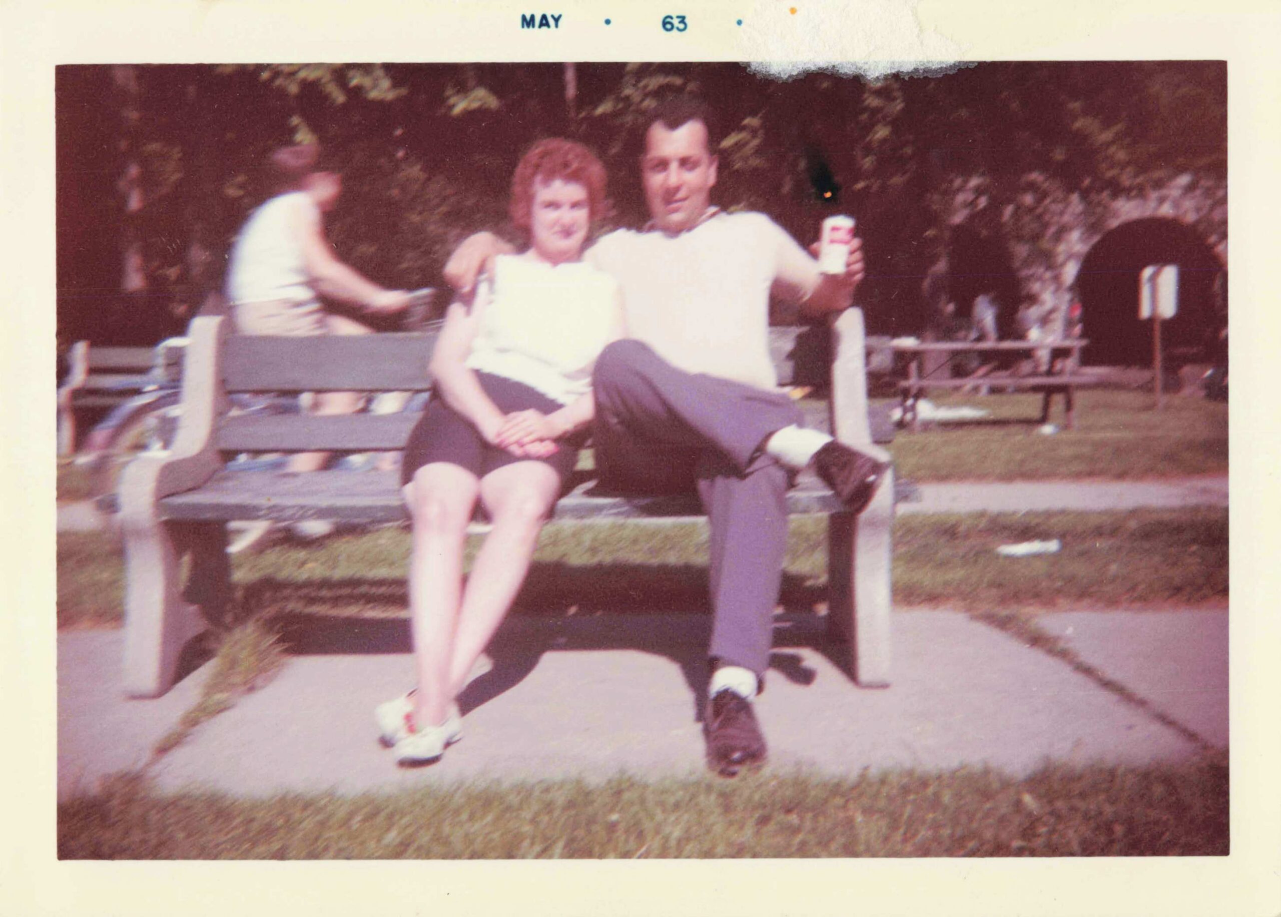 Two people sit close together on a park bench, the man has his arm around the woman and holds a can toward the camera in a casual outdoor scene.