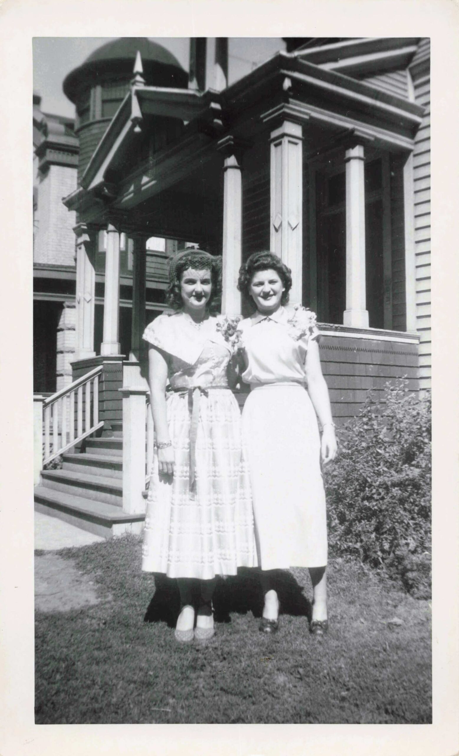 Two women in light, vintage dresses posing on a front lawn in front of a large wooden house with columns and a porch.