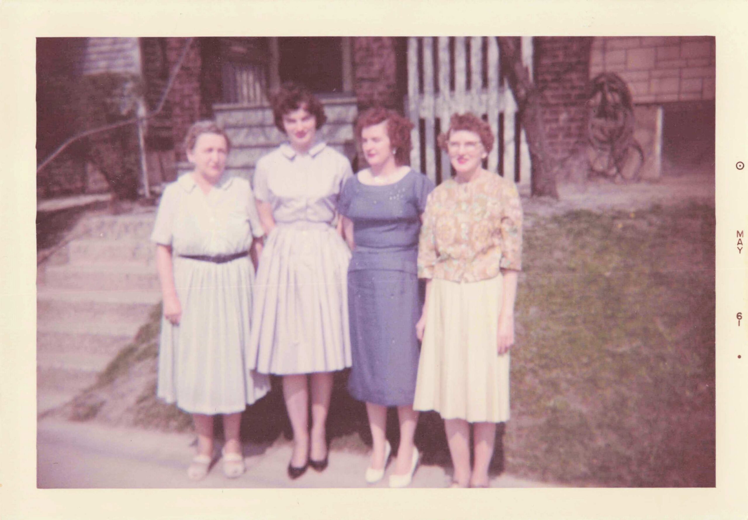 Four women in 1950s dresses posing together outdoors in front of a brick building with stone steps.