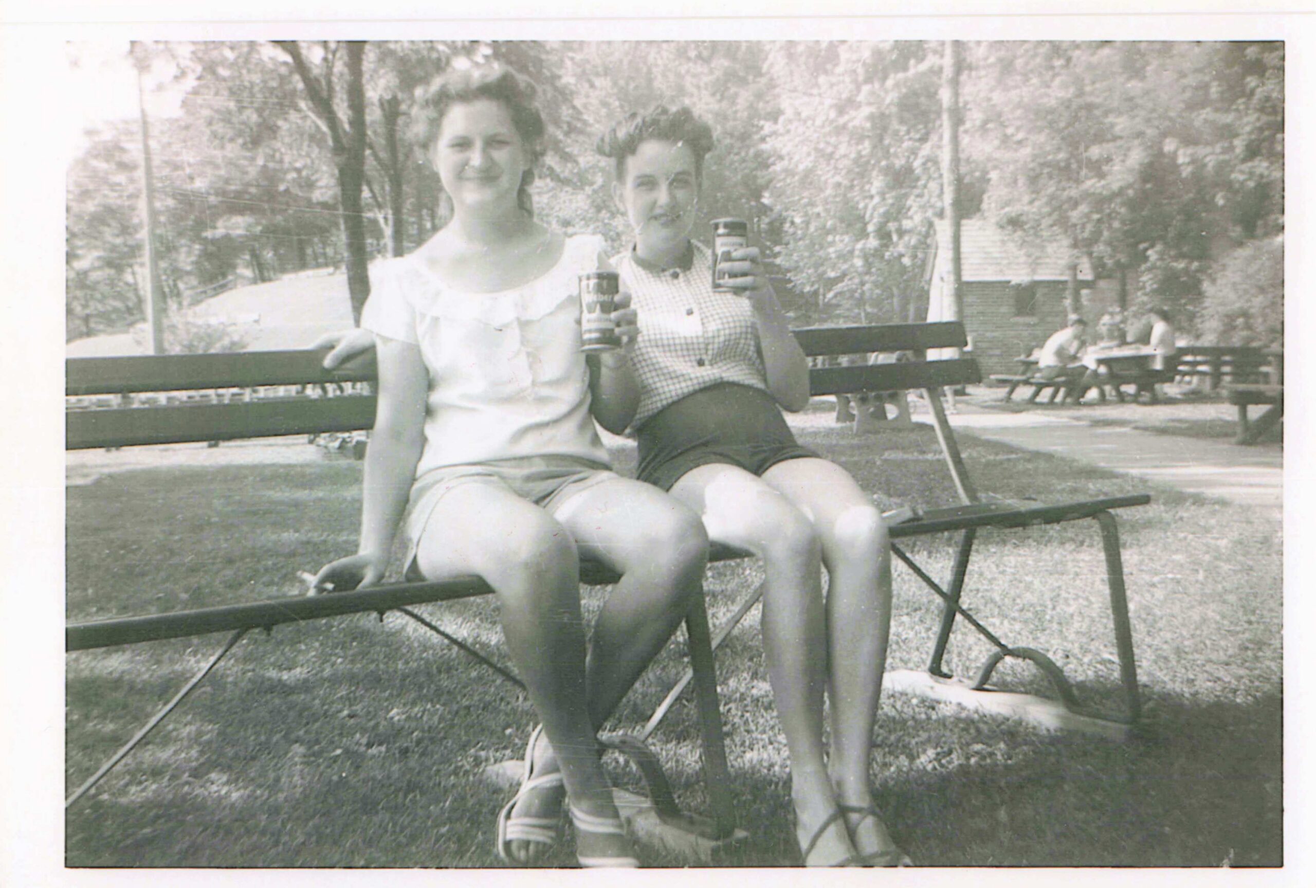 Two women sit on a park bench, smiling, each holding a drink can.