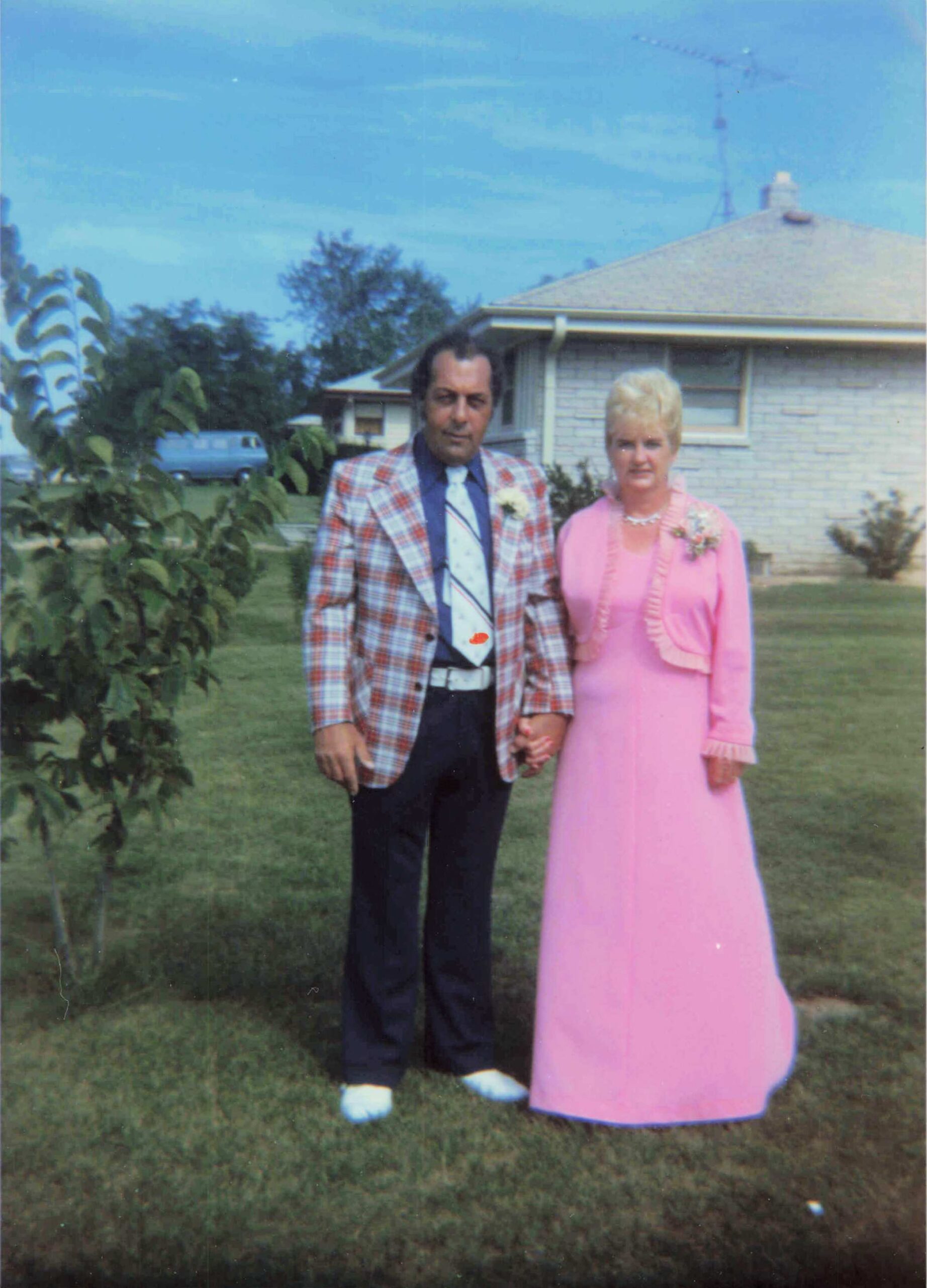 Man in a plaid suit and a woman in a pink formal dress stand hand in hand in a suburban front yard.