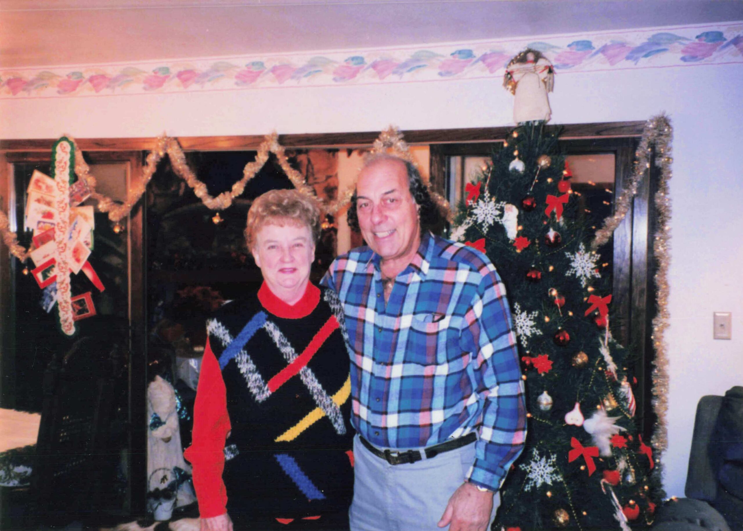 Smiling older couple posing indoors in front of a decorated Christmas tree with garlands and lights in the background.