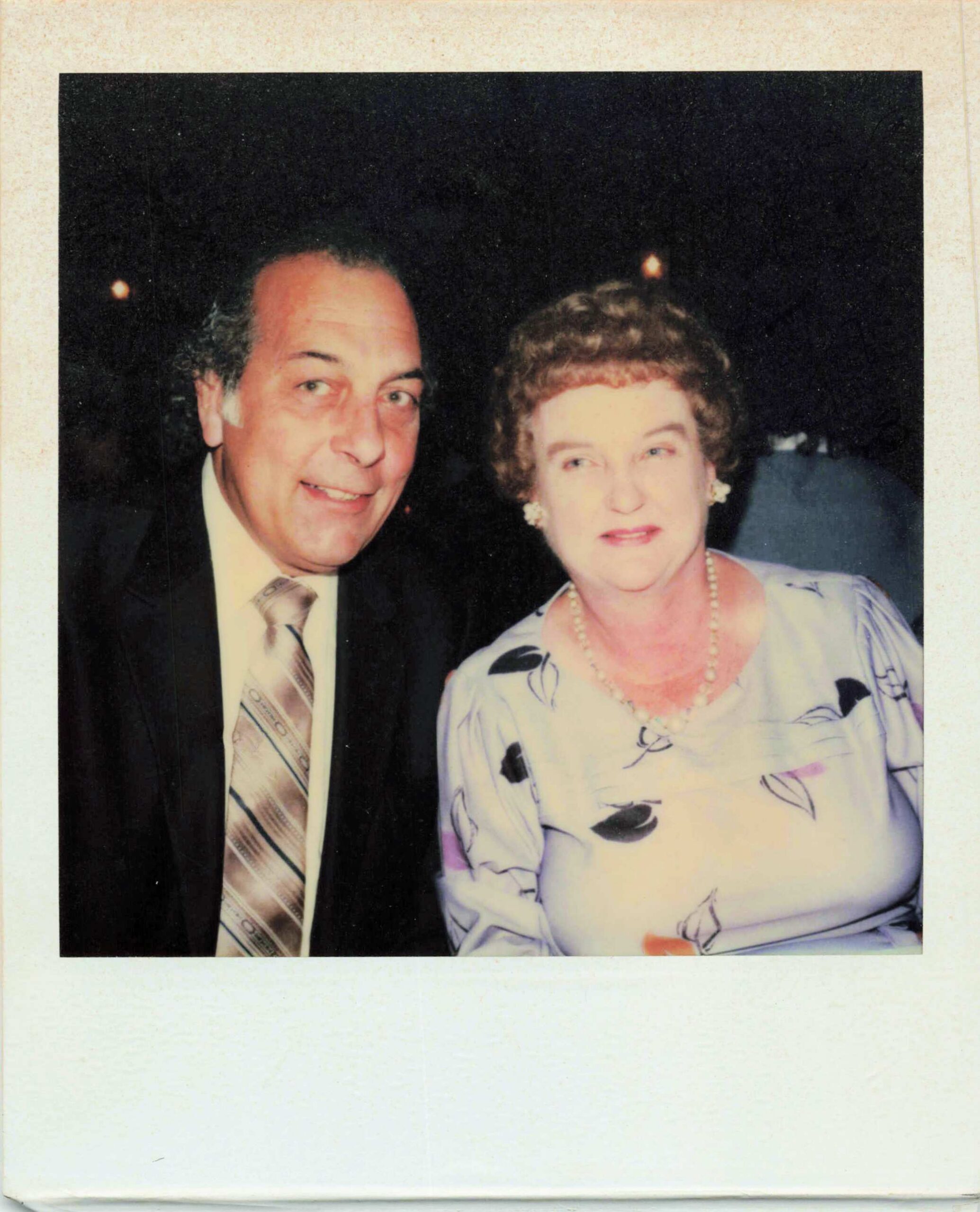Smiling older couple pose for a photo at night: man in a suit and tie, woman in light dress with a pearl necklace against a dark background.
