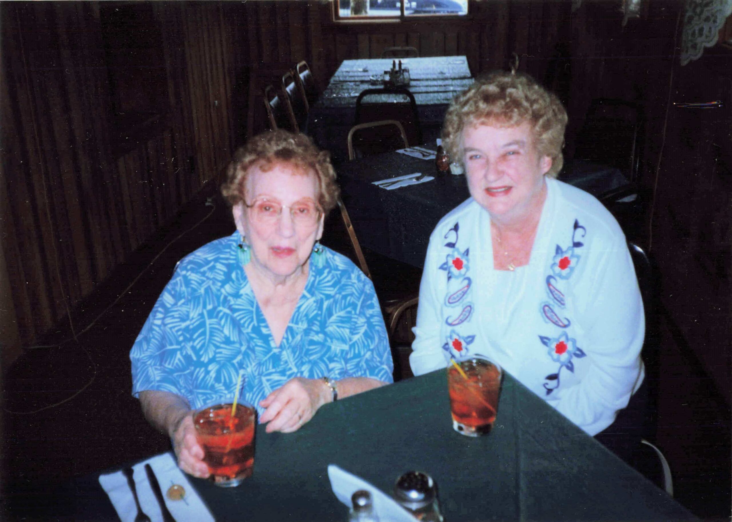 Two elderly women seated at a table with red iced drinks, wearing blue patterned tops, in a dim restaurant setting.