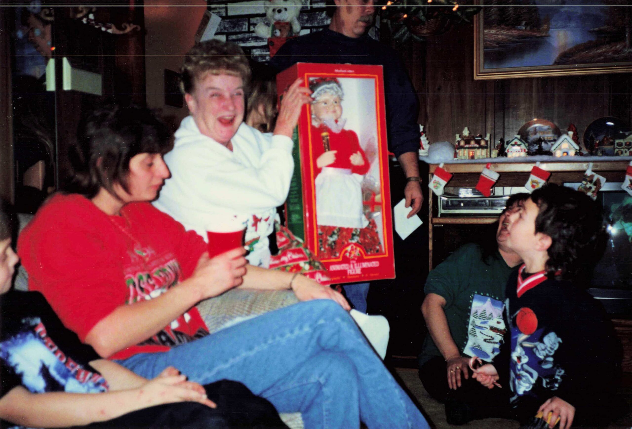 People gathered in a living room celebrating Christmas; a woman in red laughs as someone holds a boxed 'imaginative' doll gift with a girl in a red outfit on the box, others smiling nearby.