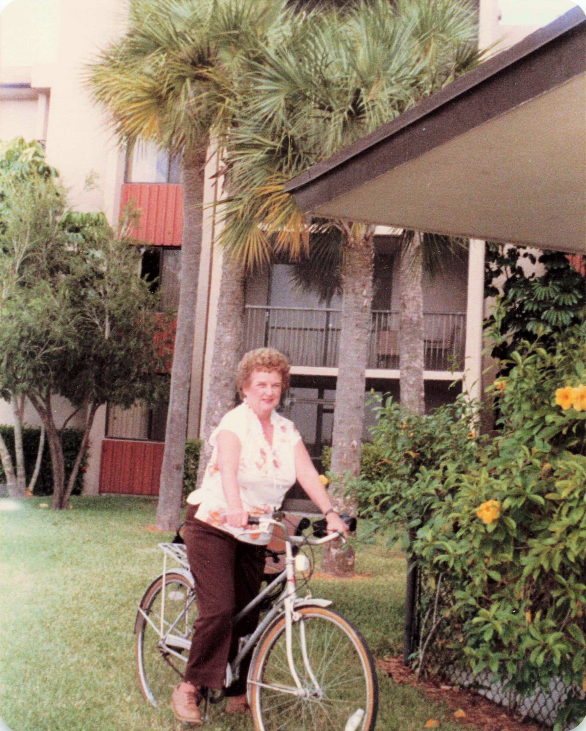 Woman wearing a floral shirt rides a white bicycle in a tropical garden with palm trees in the background, near a beige building.