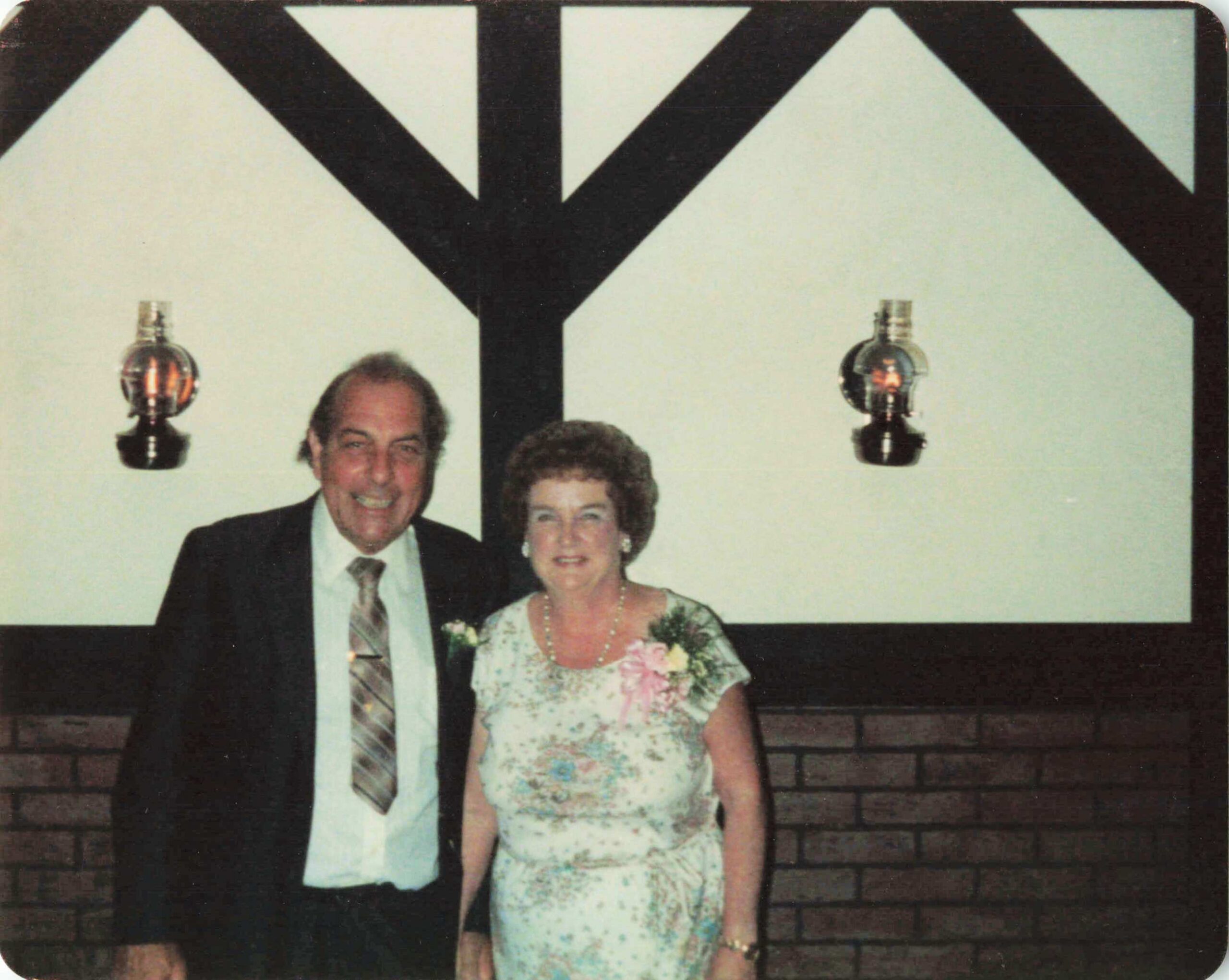 Smiling elderly couple in formal dress posing together at a wedding reception in front of a large black decorative backdrop resemble a Y shape.