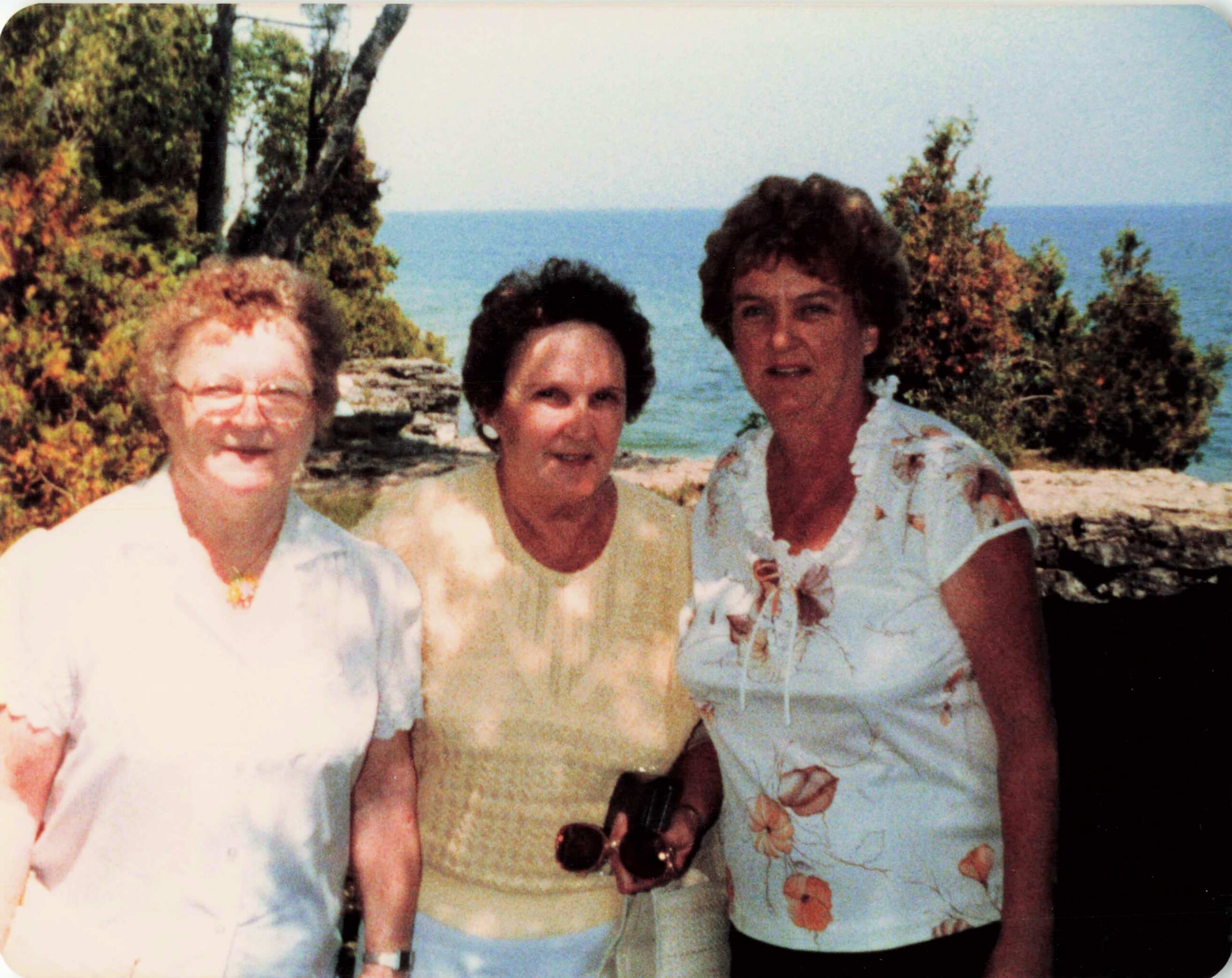 Three adults posing outdoors on a sunny beach with the sea and trees in the background, wearing light summer clothing.
