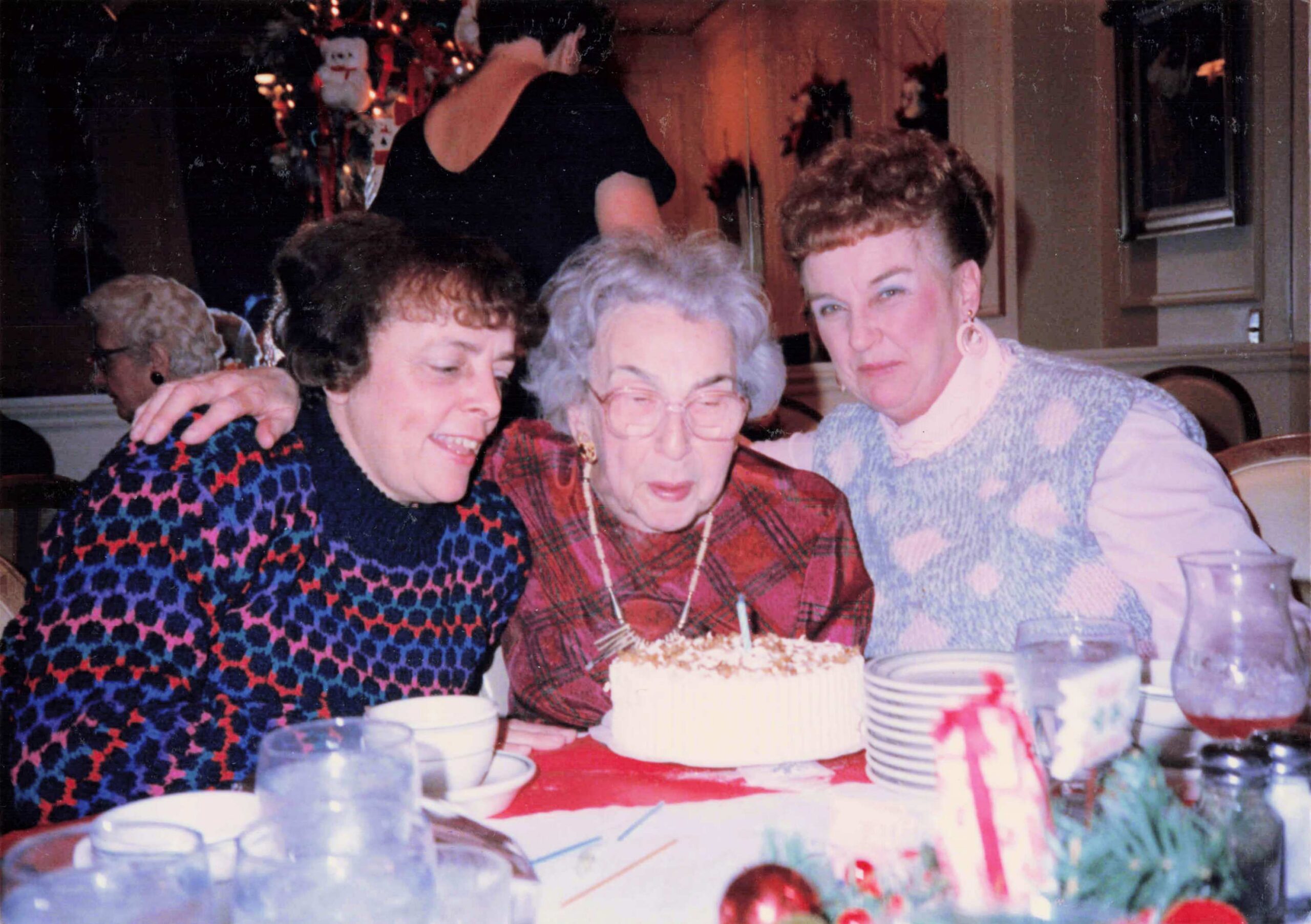 Three women pose together at a birthday party, smiling with a cake and candles in front of them, festive decorations in the background.