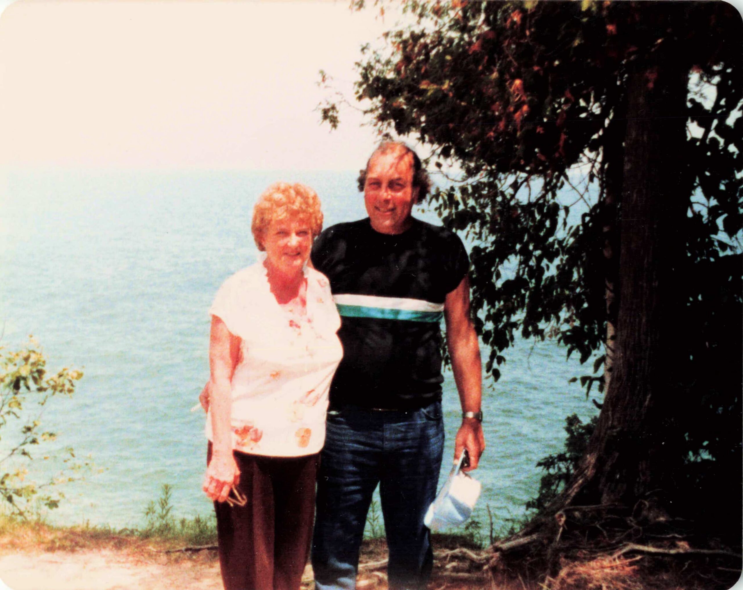 Two people standing together by a lake with a tree in the background, smiling at the camera.