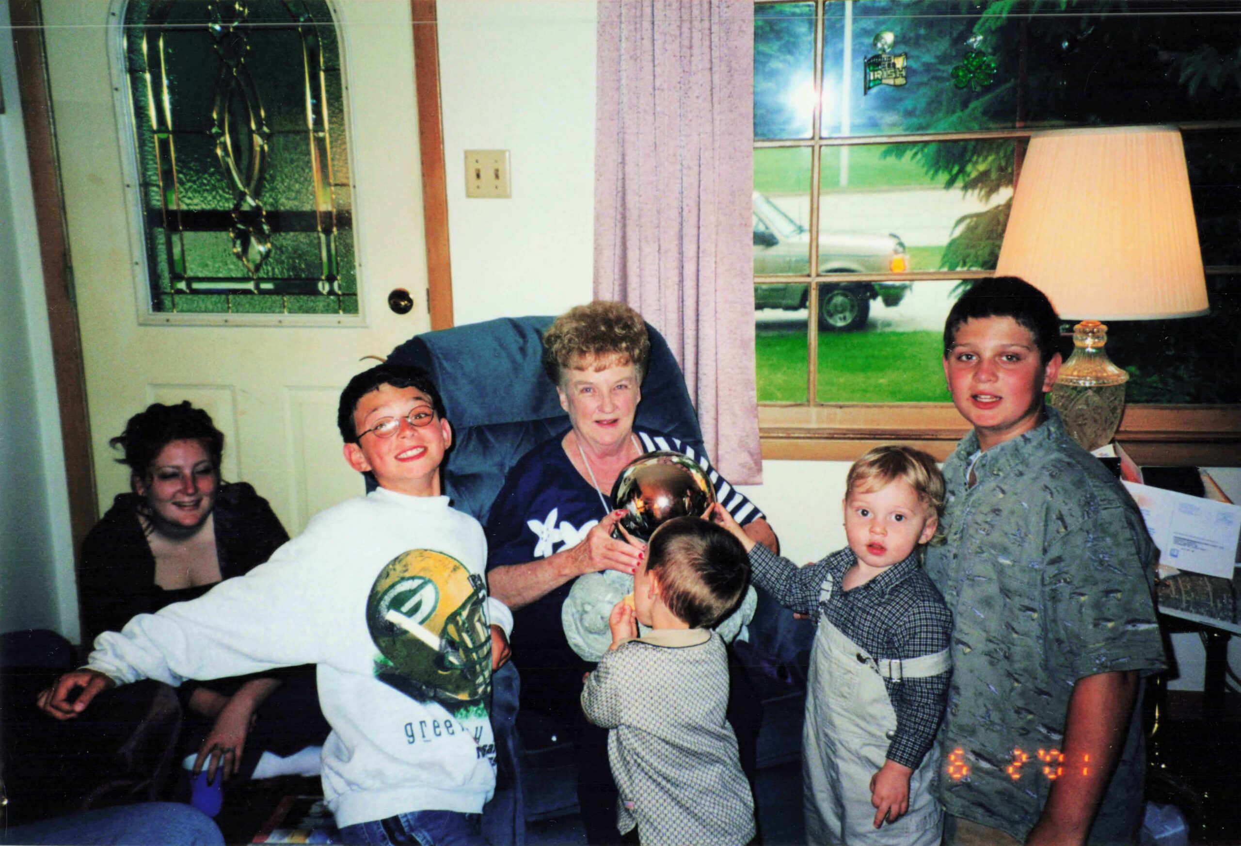 Family group gathered in a living room, several children and an older woman posing around a shiny metallic ball held by a child, with a window, pink curtains, and a lamp in the background.