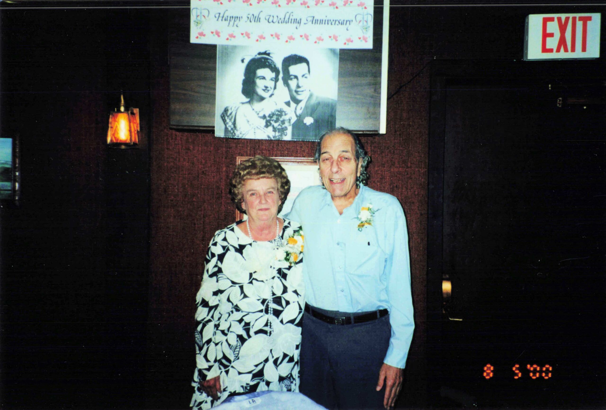 Two older adults posing together at a 50th wedding anniversary party, with a black-and-white wedding photo and a floral banner behind them.
