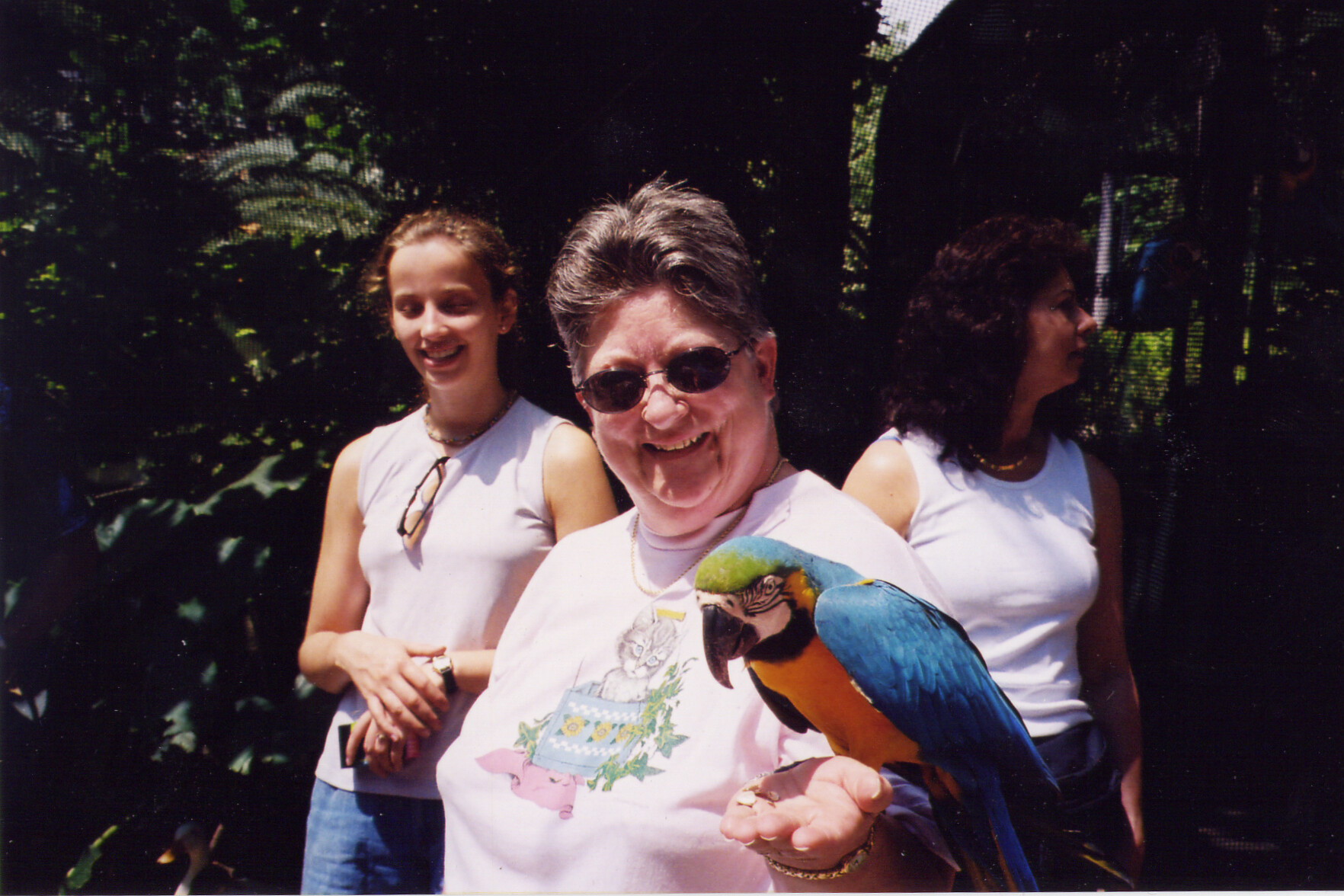 Smiling woman with sunglasses holding a colorful parrot on her arm, with two other women in white tops in a sunny outdoor setting behind her.