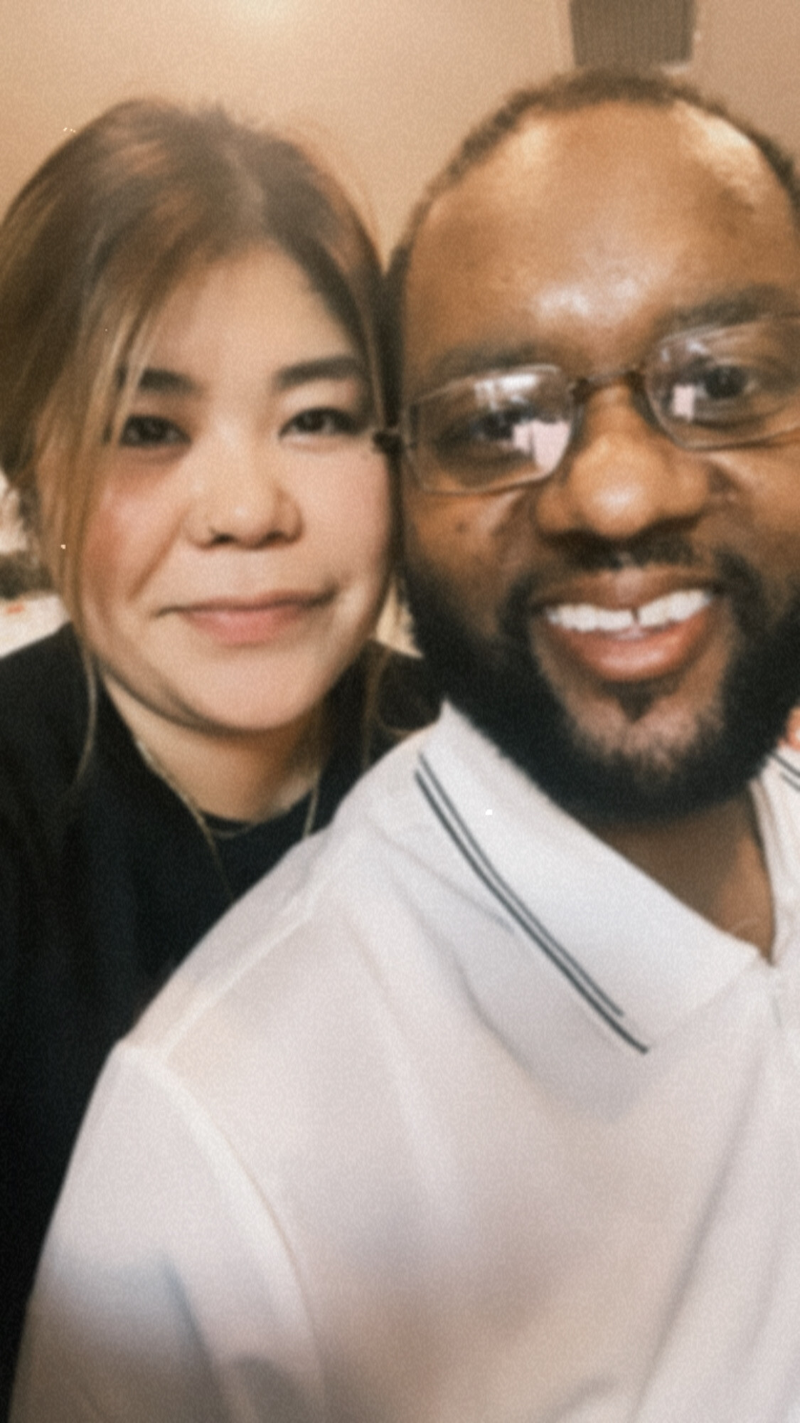 Close-up of a smiling couple taking a selfie, woman on the left and man on the right wearing glasses in a warm setting.