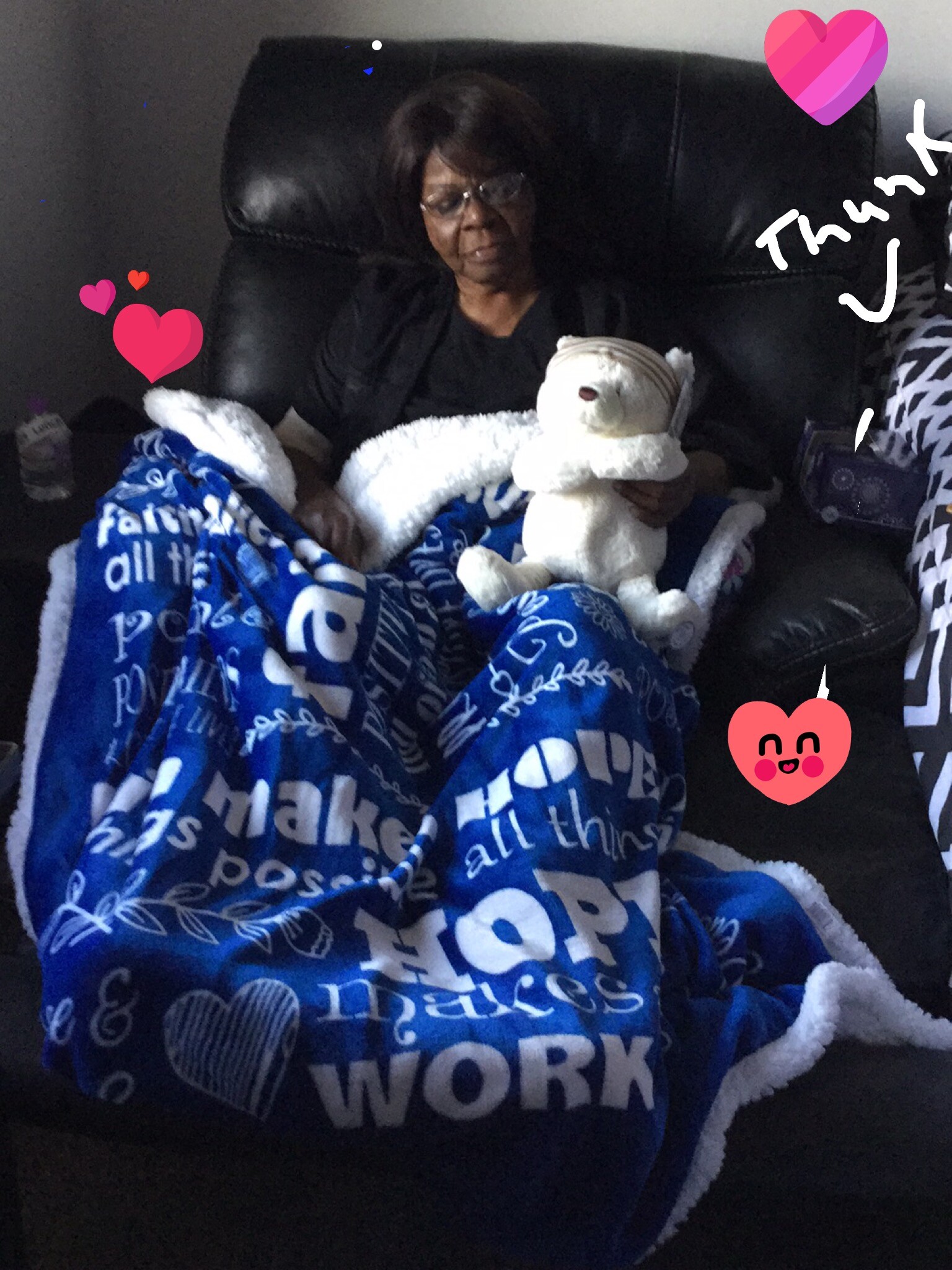 Older woman sits in a black leather chair, wrapped in a blue blanket, holding a white stuffed bear in her lap.