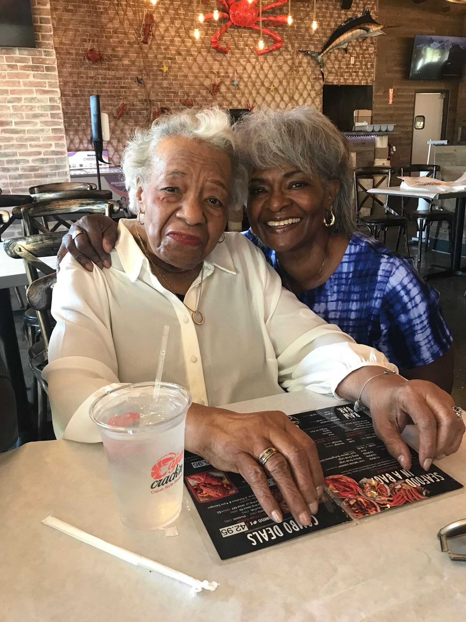Two smiling women sit at a restaurant table, one in a white blouse and the other in a blue patterned top, with a drink and a menu in front of them.