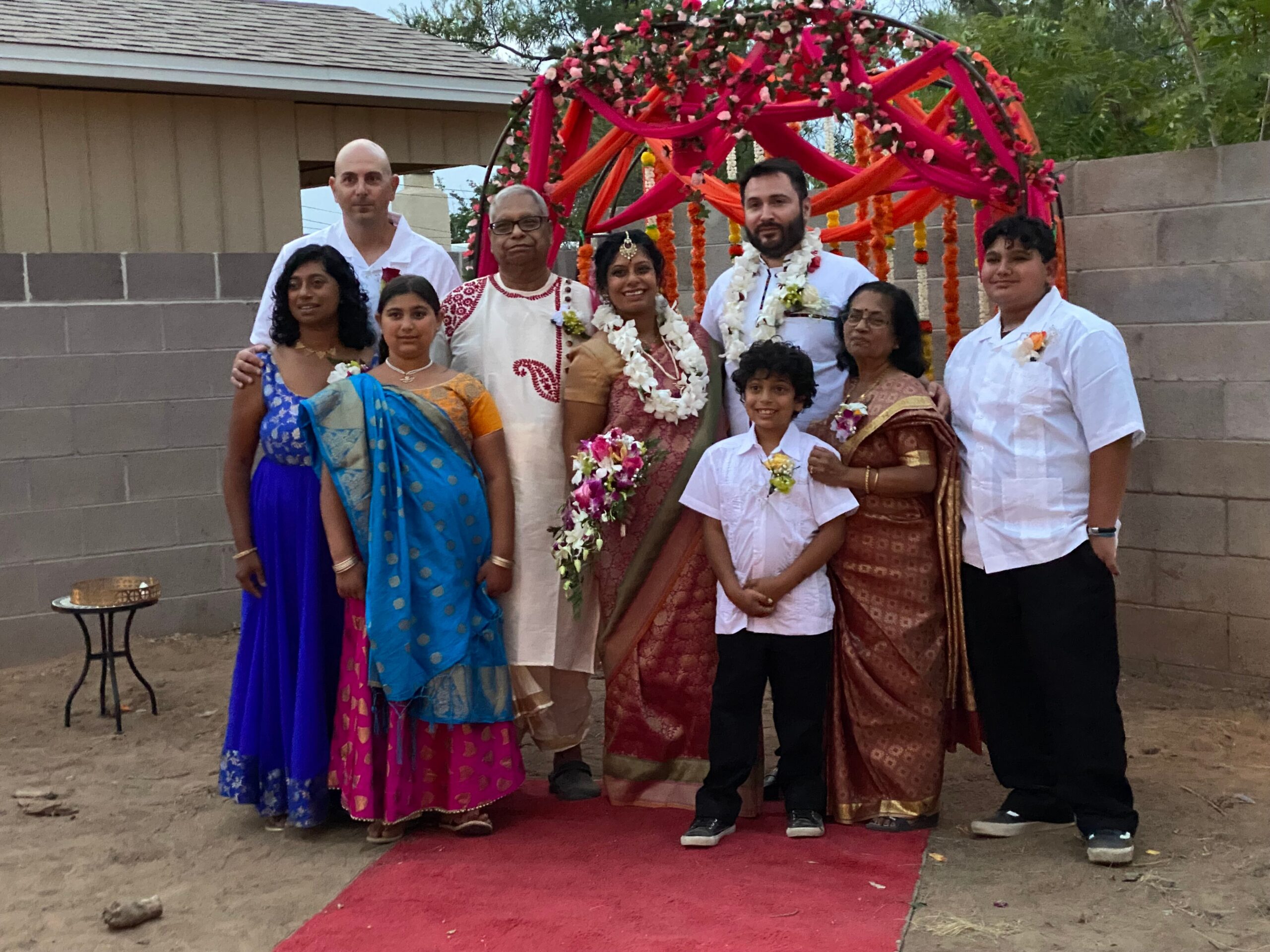 A wedding party outdoors with a couple in traditional Indian attire and family members posing on a red carpet under a flower arch.