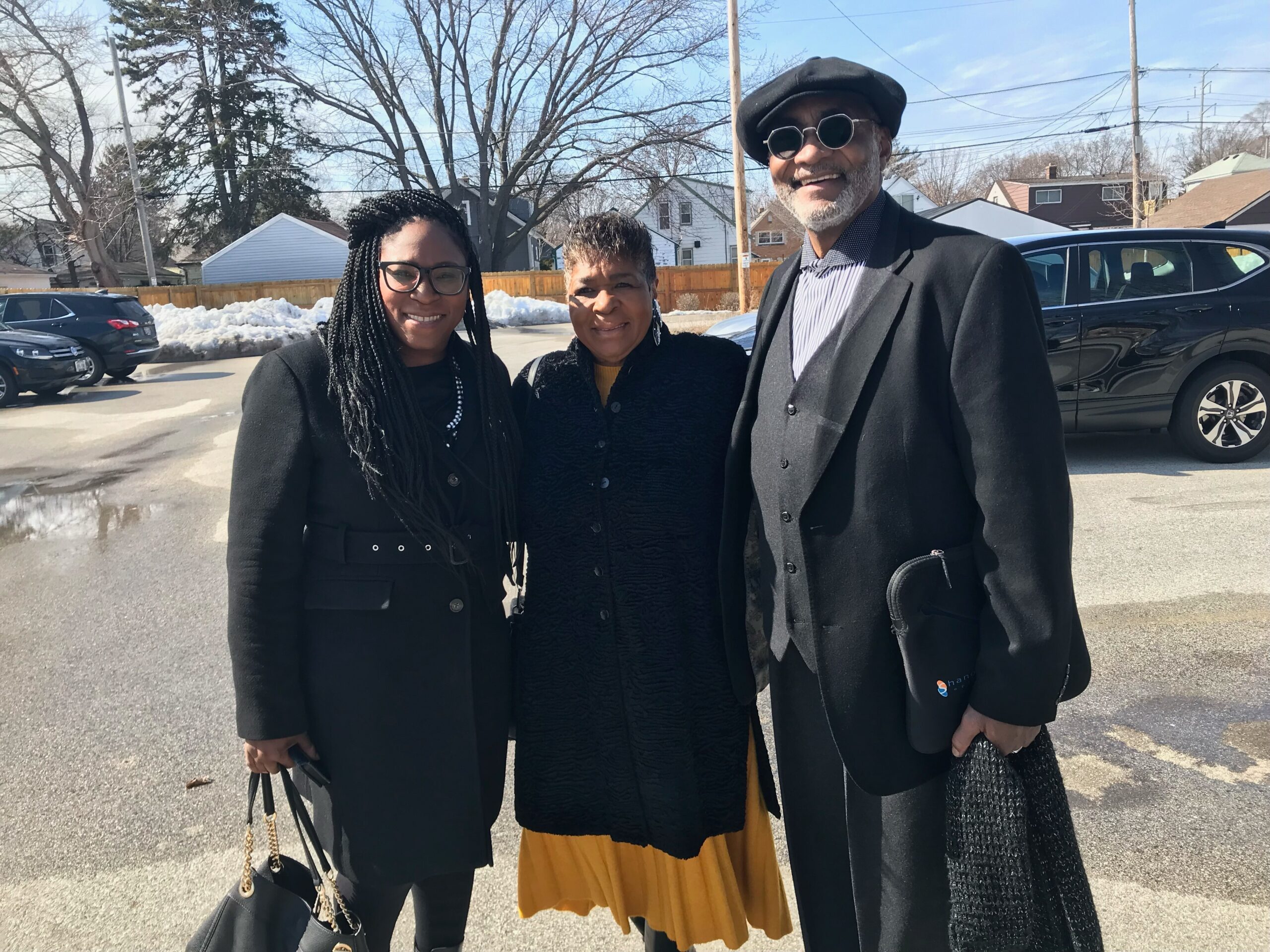 Three adults posing together outside on a sunny winter day, all wearing dark coats and smiling at the camera.