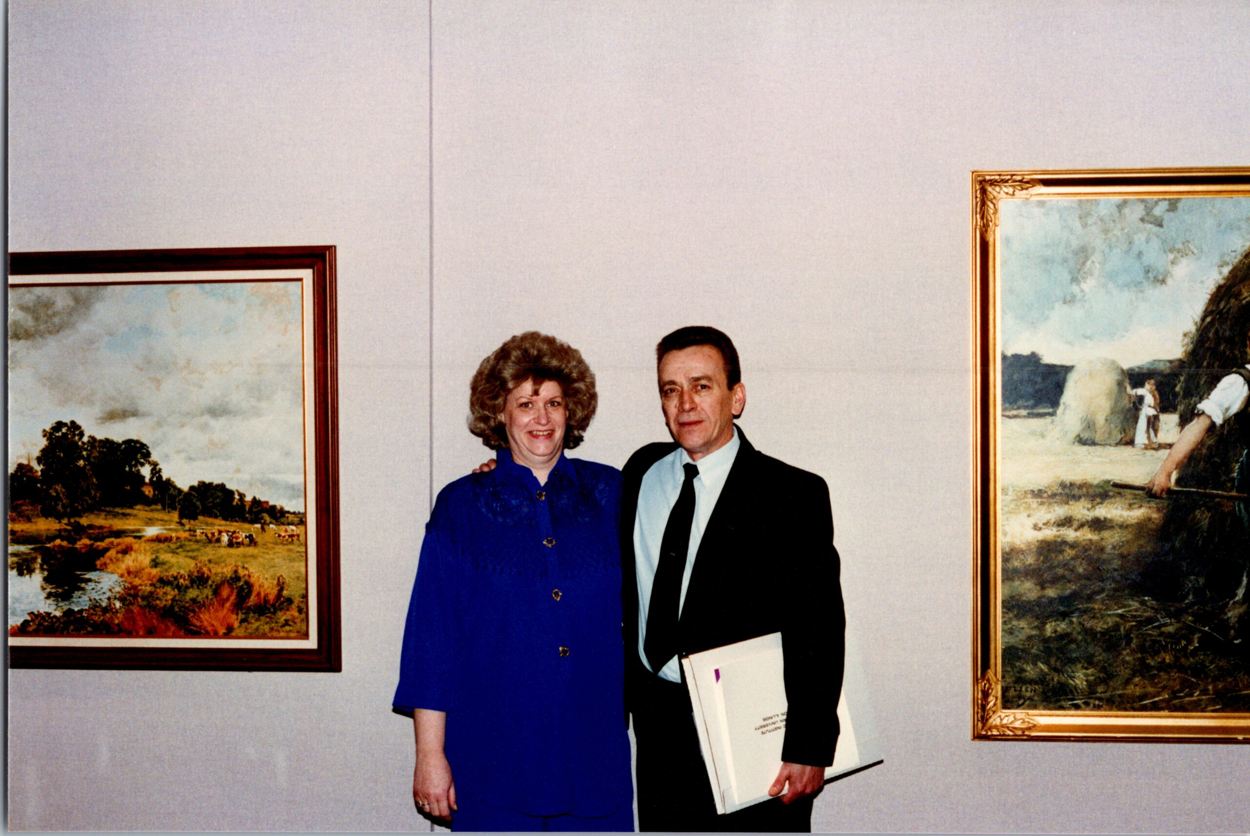 Two adults stand together in an art gallery between framed landscape paintings on the wall behind them.