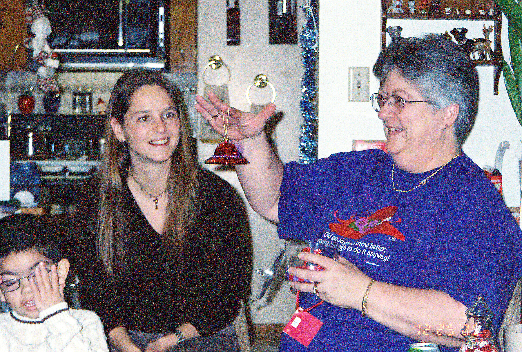 Woman in a dark top smiles as an older woman in a purple shirt presents a red bell ornament at a family gathering in a kitchen.