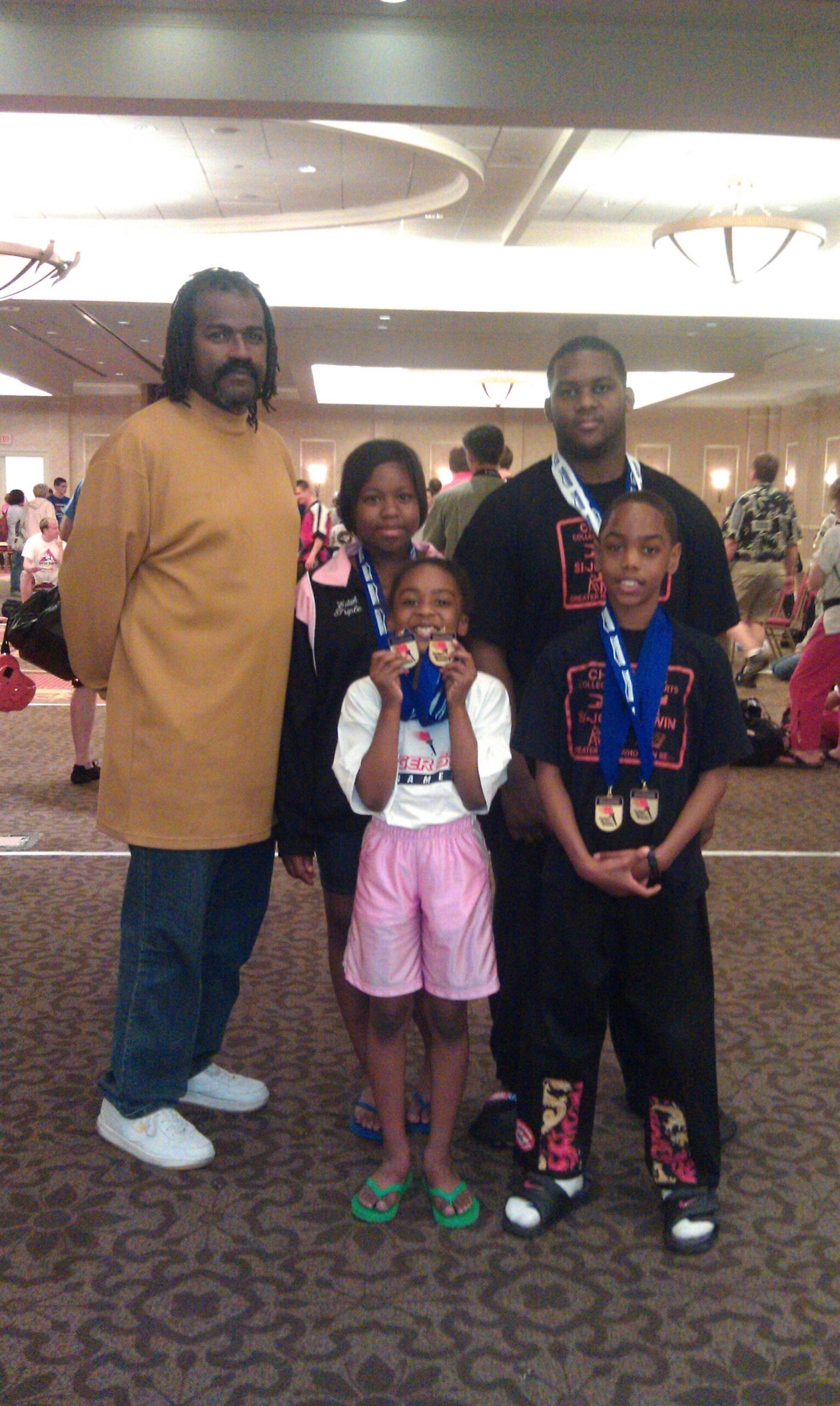Group of five people posing for a photo at an indoor awards event; two adults (one tall man in tan shirt) with three children wearing medals.