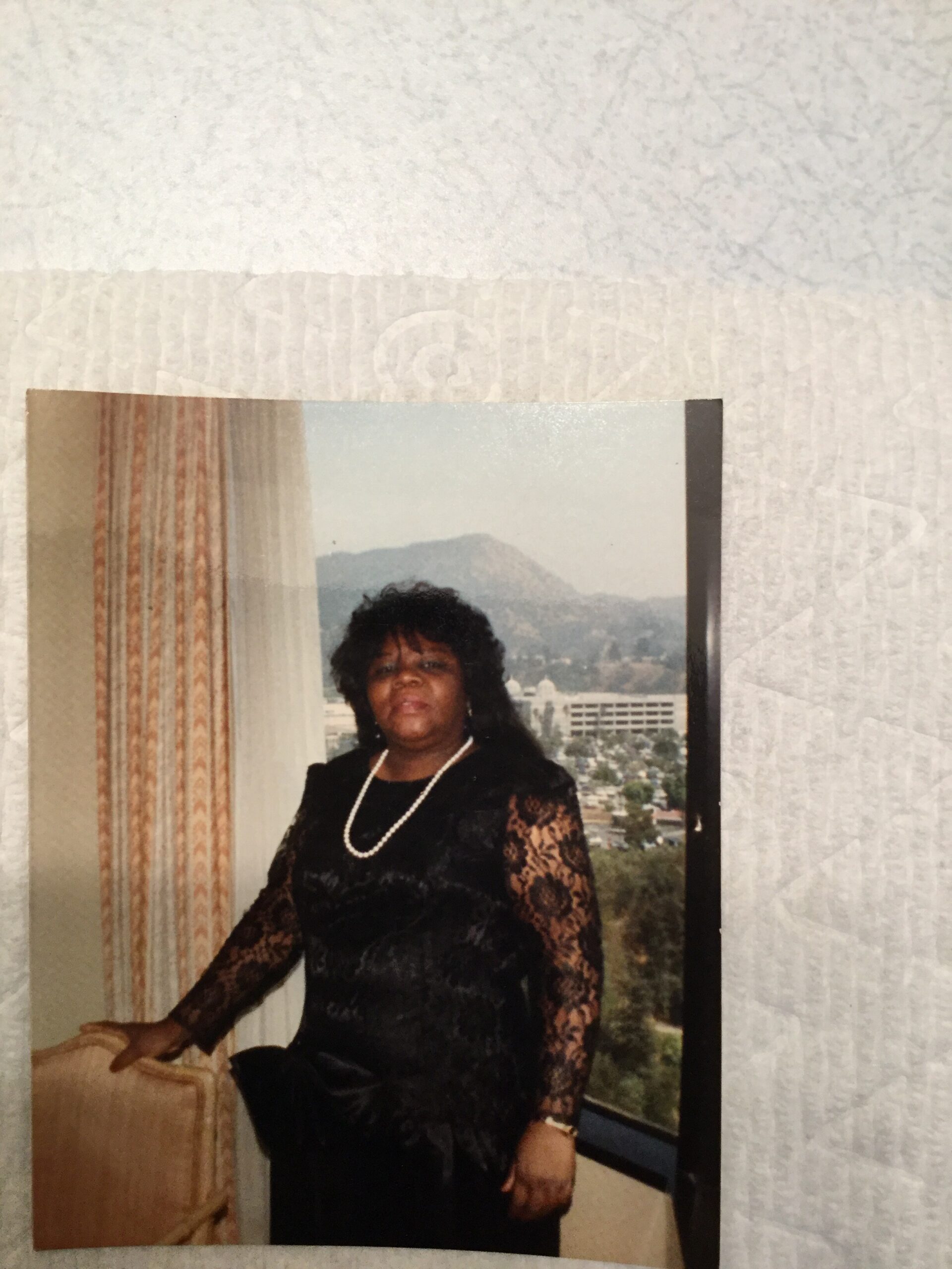 Woman in a black lace dress with a pearl necklace standing near a window overlooking a cityscape, indoors.