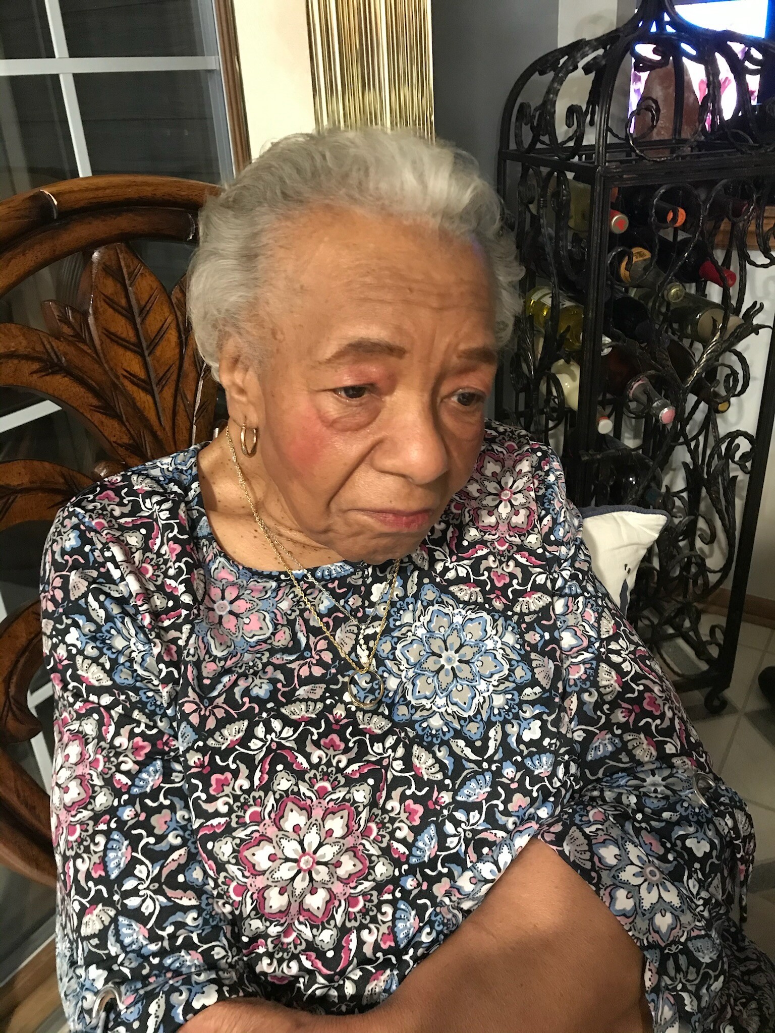 Close-up of an elderly woman with short gray hair wearing a floral blouse, seated indoors near a decorative wine rack.