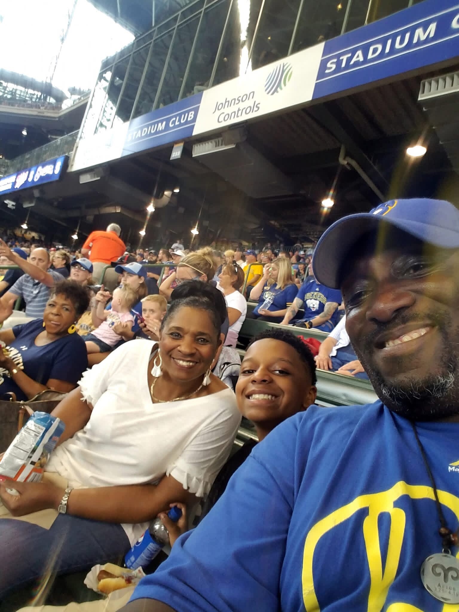 Smiling spectators in stadium seats taking a selfie, with a blue Stadium Club sign and Johnson Controls banner overhead.
